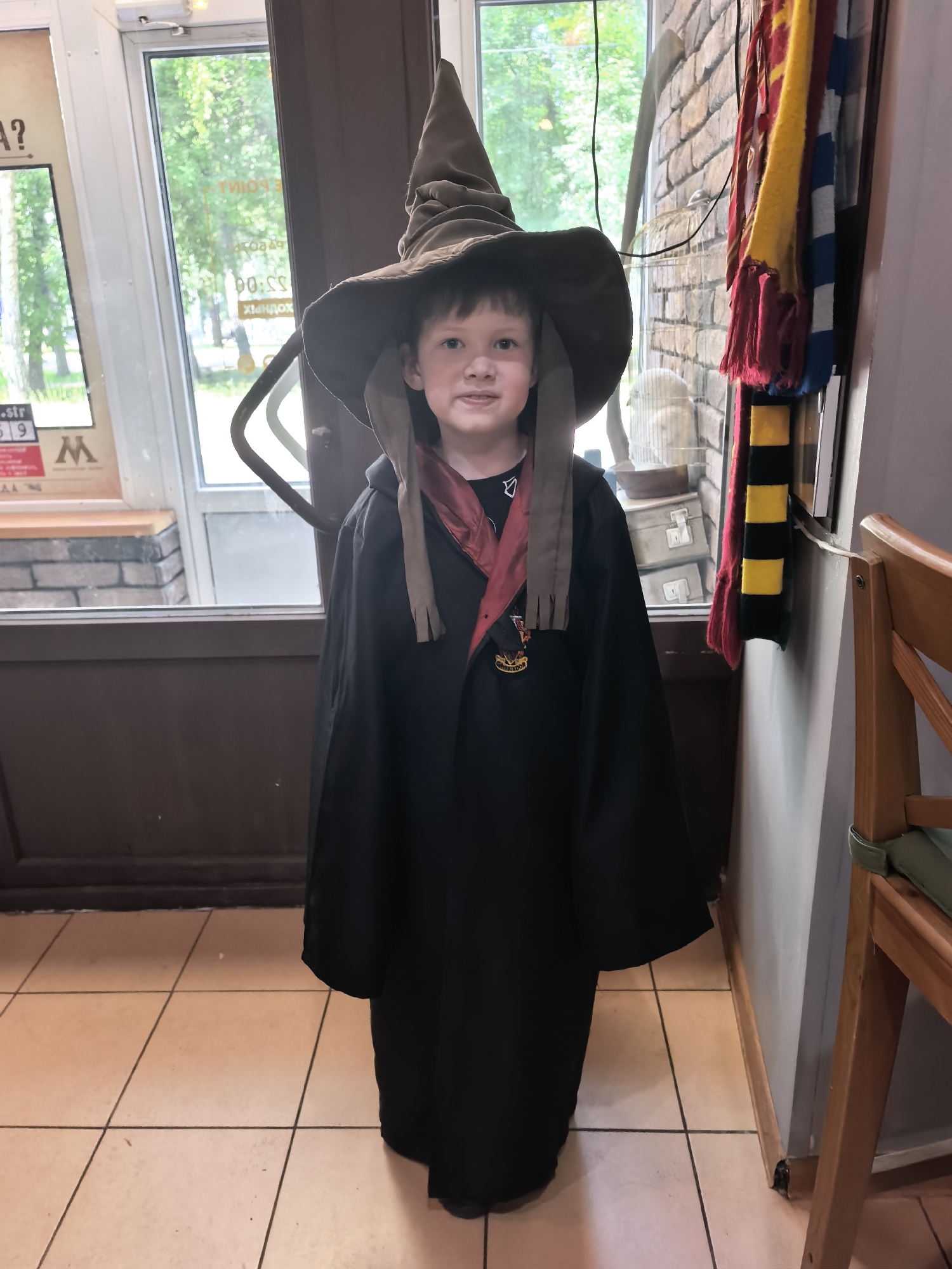 A young boy is posing for a picture, dressed in a wizard costume reminiscent of Harry Potter. He wears a large, pointed hat and a black robe over a dark shirt. A red and gold scarf hangs around his neck, and a Hogwarts crest badge is pinned to his robe. He is smiling faintly, looking directly at the camera with a playful expression.
The background suggests an indoor setting, possibly a shop or event space, with elements hinting at a theme. To the left, a window looks out onto a green, leafy exterior, indicating it's daytime and likely pleasant weather. There are posters on the window, one with the letter "M" and some text. A brick wall texture is visible on the right, and colorful scarves, including one with black and yellow stripes, are hanging nearby. A wooden chair is partially visible on the far right. The floor is tiled with a light brown color. The overall mood is cheerful and whimsical, likely capturing a moment of fun or a special occasion.