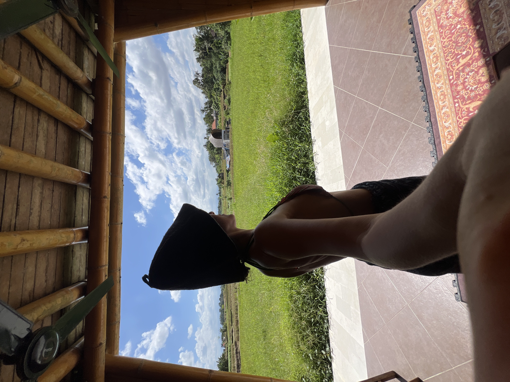 A person wearing a black hat, dark top, and dark bottoms is standing on a tiled surface inside what appears to be a bamboo structure. The person is facing a vista of a green rice field, possibly in the Ubud District of Indonesia. The sky is blue with puffy white clouds. A patterned rug is partially visible on the tile floor.