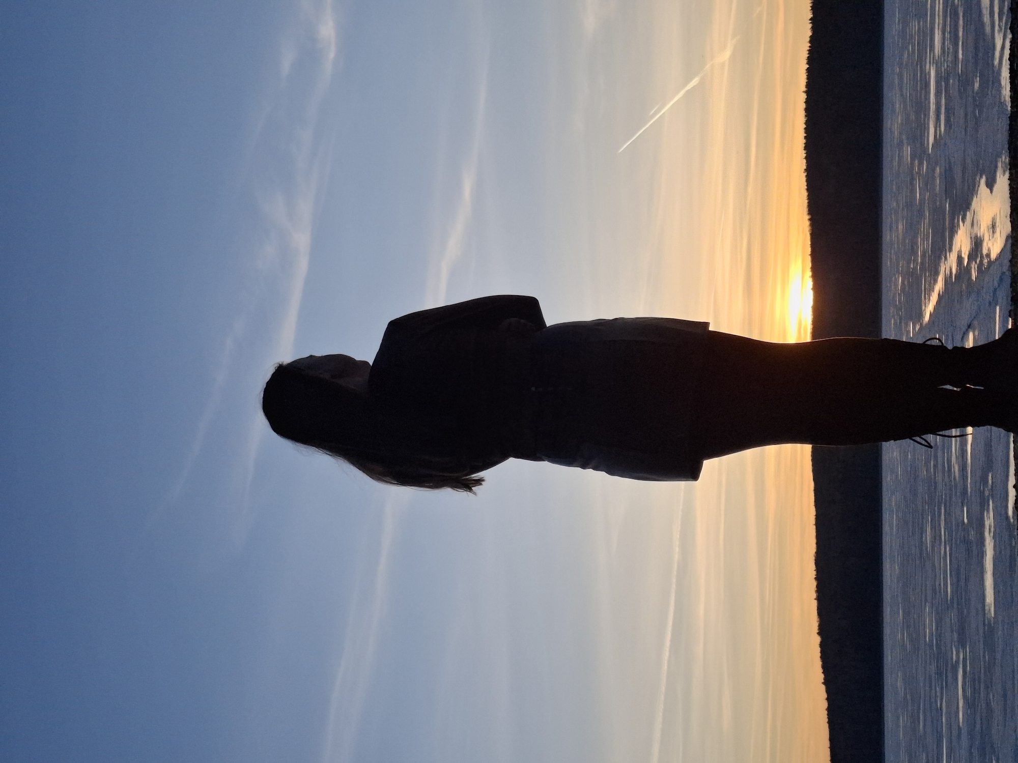 The image captures a solitary figure silhouetted against a dramatic sunset over a body of water, likely in Chaikovsky, Russia. The person, appearing to be a woman with long hair, stands facing the horizon, with her arms bent, possibly folded or holding something. She is dressed in a dark, indistinct jacket or coat, her form completely blacked out by the intense backlighting.
The sky dominates the upper portion of the scene, transitioning from a clear, deep blue on the left to a brilliant orange and yellow glow on the right, where the sun is actively setting on the horizon. A bright glare emanates from the sun. Sparse, wispy clouds are visible, along with a distinct white airplane contrail stretching across the middle of the sky.
A dark, rugged line separates the sky from the water, suggesting a distant treeline or landmass. Below this horizon line, a body of water reflects the golden hues of the sunset. The reflections appear as shimmering, elongated patterns on the water's surface, indicating either ripples or possibly partially frozen, textured water, given the location context.
The scene depicts a peaceful moment at sunset, conveying a sense of contemplation or admiration for the natural beauty. There is no visible text in the image.