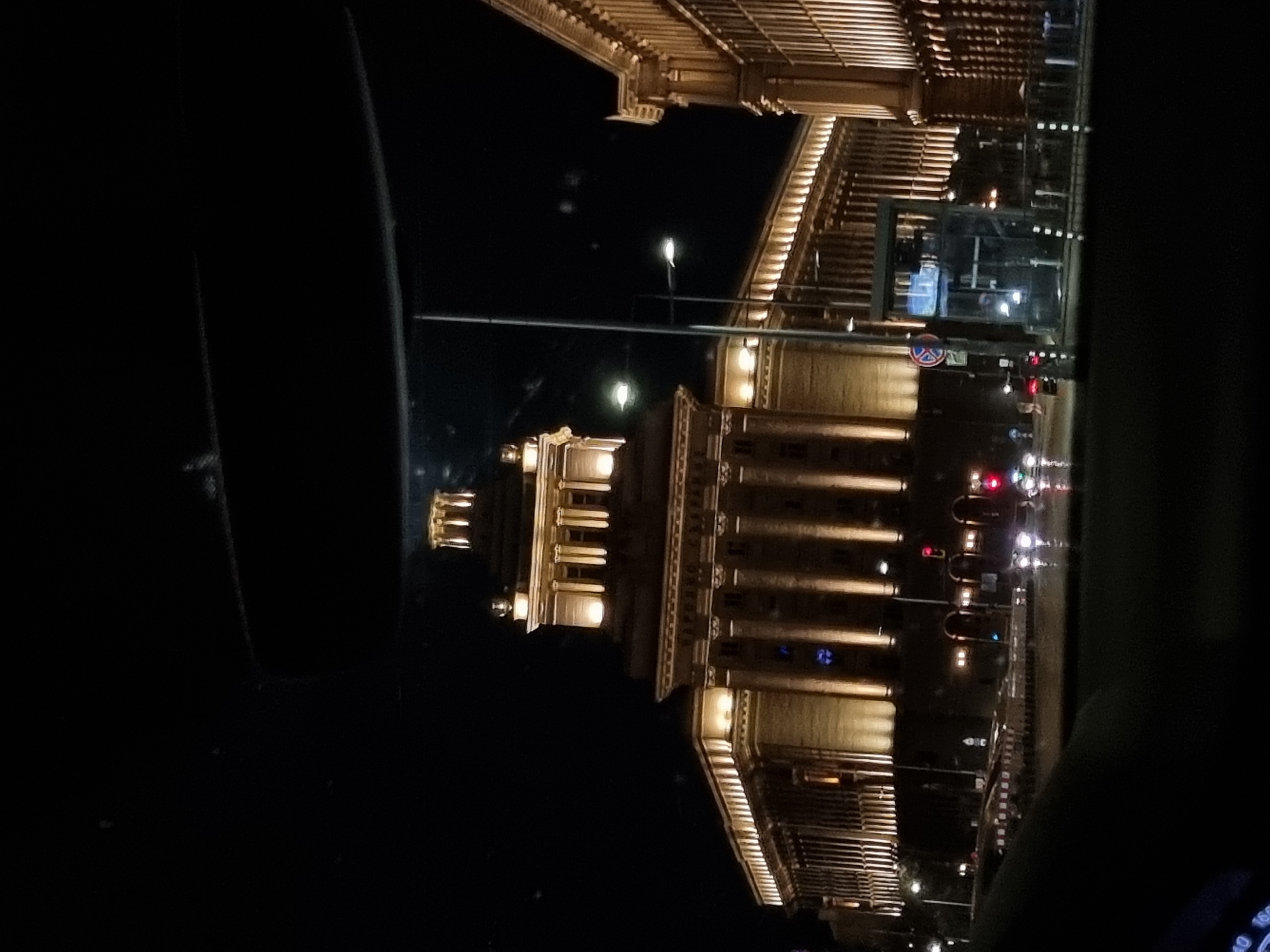 The image shows the Palace of Justice in Sofia, Bulgaria at night. The building is brightly lit, highlighting its neoclassical architecture, including columns and architectural details. Street lights and traffic lights are visible in the foreground. The image appears to have been taken from inside a vehicle, as part of the dashboard and the window frame are visible in the darkness of the car interior.