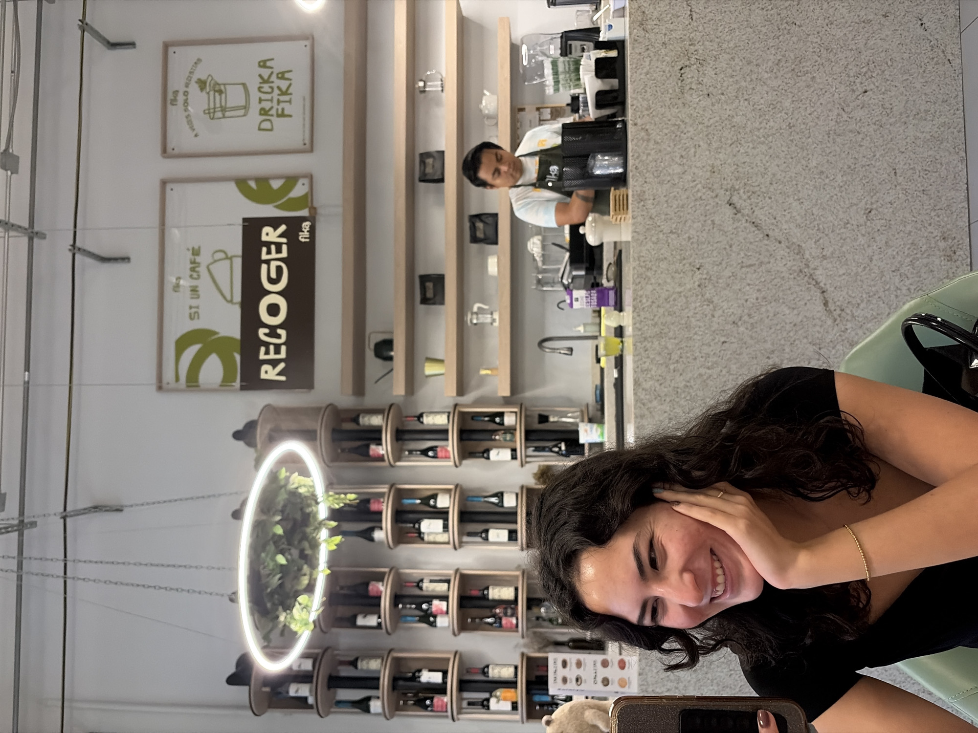 A woman is sitting at a cafe, holding her phone, with her hand on her face, smiling. The cafe has a coffee bar with a barista behind it, shelves of wine bottles, and signs that say "DRIČKA FIKA" and "RECOGER fika". The location is in the city of Puebla, Mexico.
