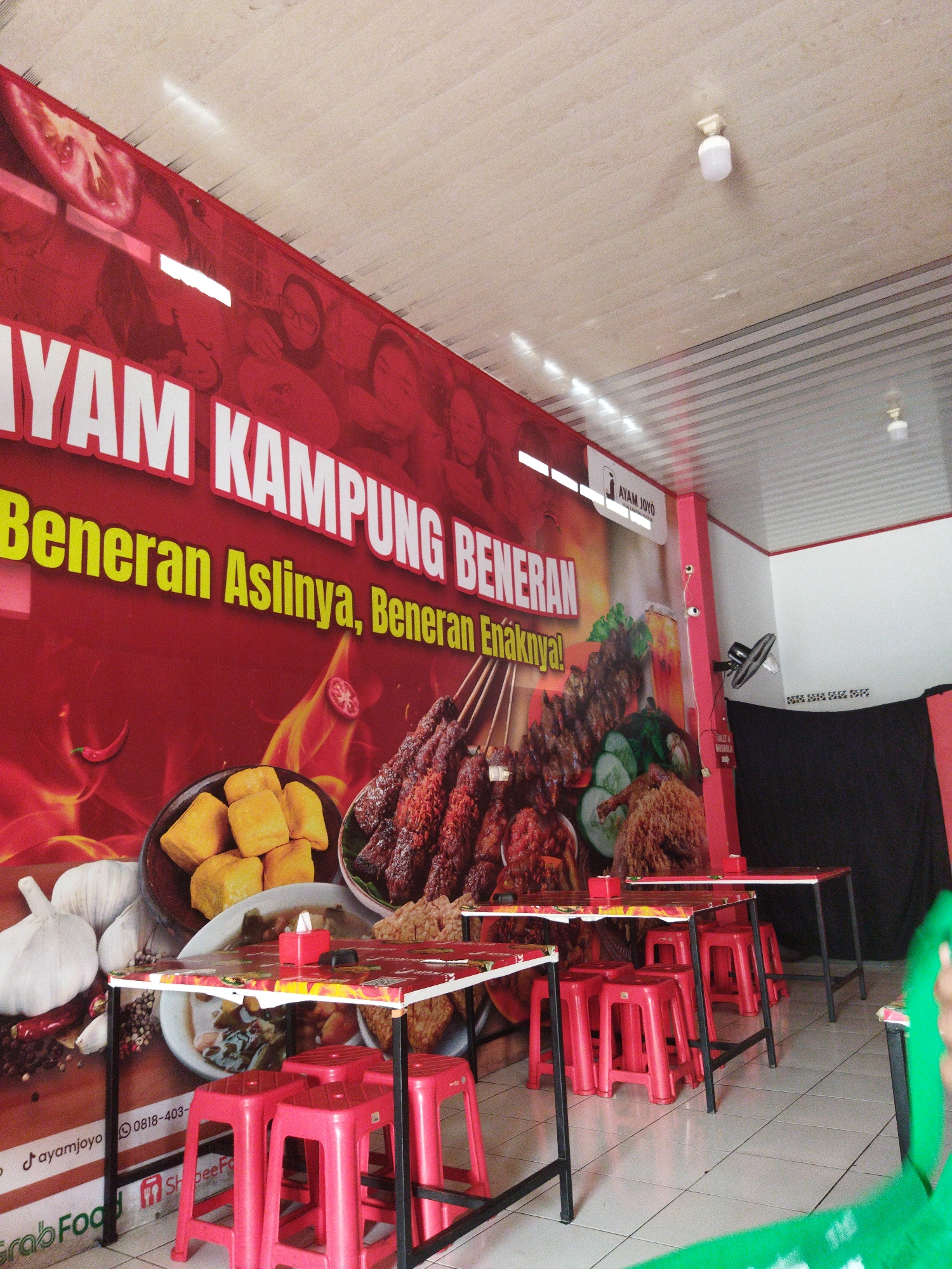 The image captures the interior of a casual dining establishment, likely a restaurant, with a prominent red banner featuring text in white and yellow. The banner displays "AYAM KAMPUNG BENERAN" in large white letters, followed by "Benaran Aslinya, Benaran Enaknya!" in yellow. Below this, there are images of food, including fried tofu, grilled chicken skewers, and what appears to be fried chicken. The environment is simple, with white tiled floors and a suspended ceiling. Several red plastic stools are arranged around tables with white tablecloths featuring colorful food imagery. In the background, a black curtain hangs, and a black fan is mounted on a pink pillar. There are also signs and logos visible, including "AYAM JOYO" on a white sign and social media/delivery service logos such as "GrabFood" and "ShopeeFood" on the lower left. The overall impression is of an informal eatery, possibly in Tangerang, Indonesia, given the presence of local food items and delivery service logos commonly used in the region.