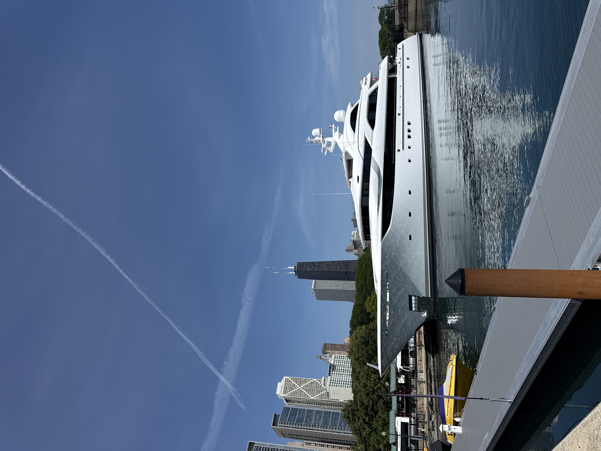 A large, white luxury yacht is docked in the harbor of Chicago, Illinois. The yacht is positioned in the foreground, taking up a significant portion of the right side of the frame. Its sleek, modern design is evident with multiple levels and large windows. The yacht is reflecting in the calm, blue water of the harbor.
In the background, the Chicago skyline is visible. Prominent buildings include the Hancock Center, identifiable by its distinctive cross-braced structure, and other modern skyscrapers. Green trees line the waterfront, separating the harbor from the urban landscape.
A gray dock with a brown wooden post extends into the foreground from the left side of the image, suggesting the viewer is on a pier. A small, bright yellow boat is tied to the dock to the left of the yacht. The sky is a clear, bright blue with a few white contrails from airplanes streaking across it. The sun is shining, creating reflections on the water and illuminating the scene. The overall impression is a bright, sunny day in a bustling urban harbor.