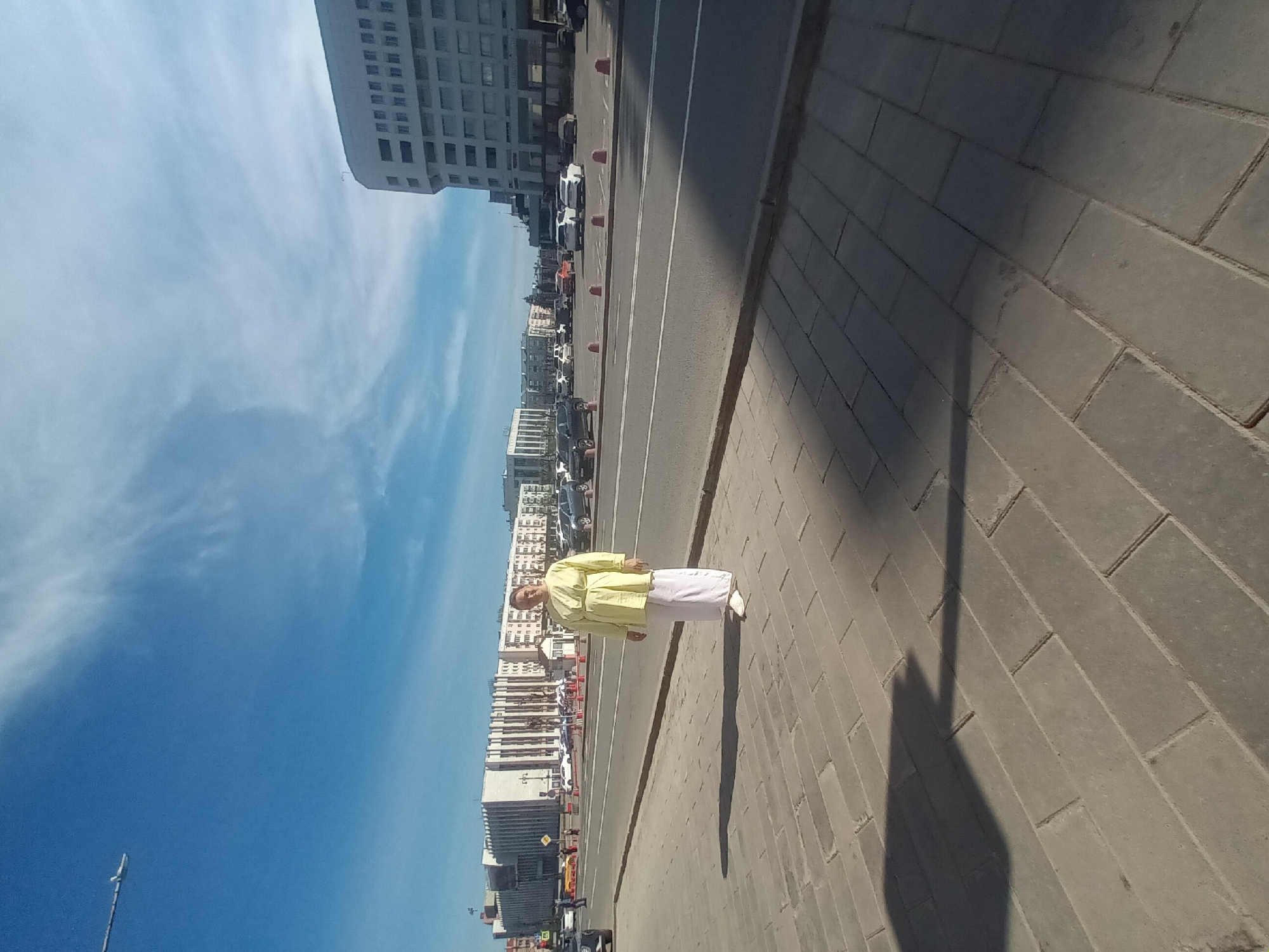 The image shows a street scene in Perm, Russia. A person wearing a light yellow shirt and white pants is standing on a brick sidewalk. Behind the person is a road with parked cars and buildings on the other side. The sky is blue with some clouds.