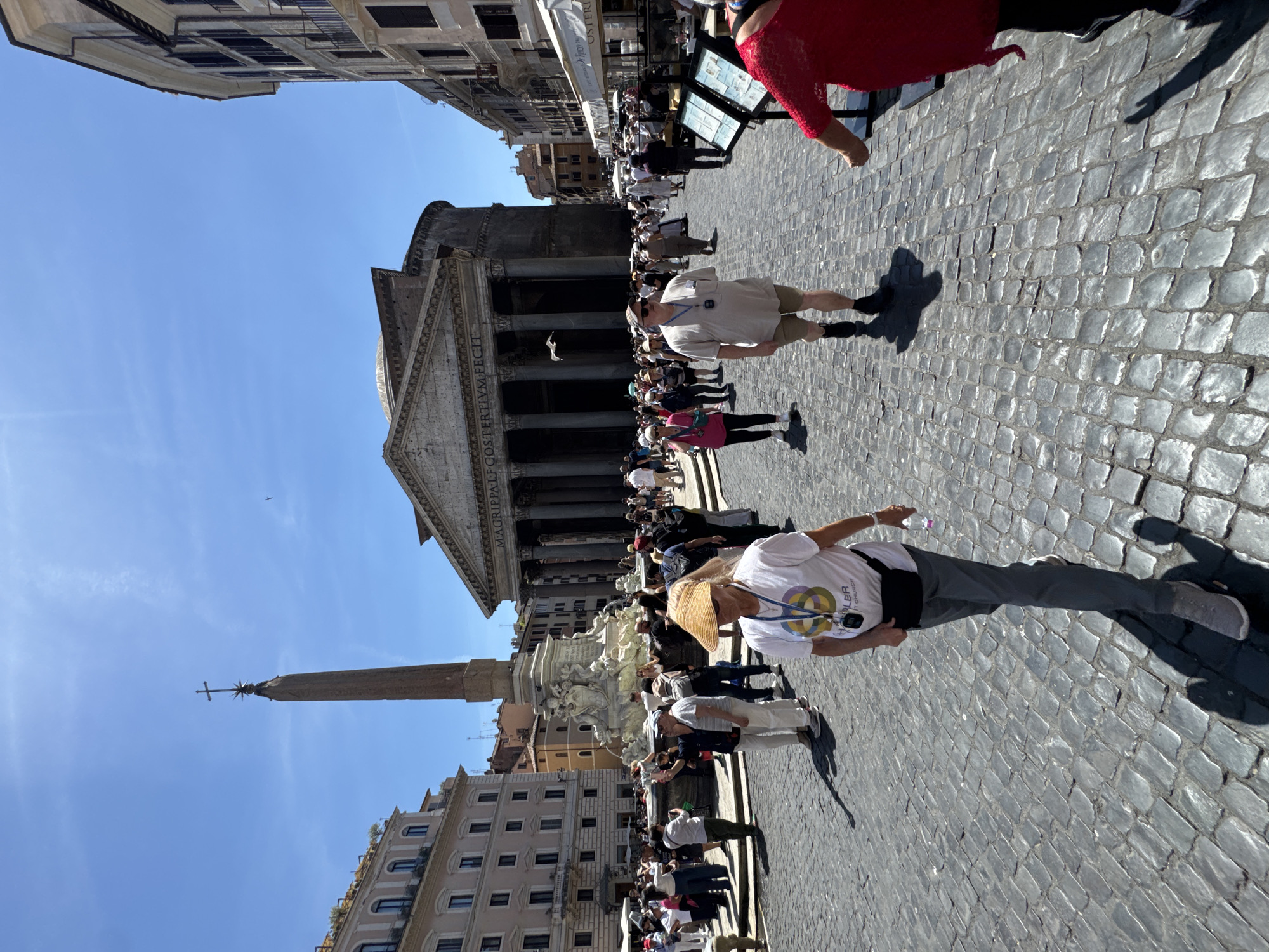 The image shows a street scene in Rome, Italy, featuring the Pantheon. The Pantheon is prominently displayed, along with an obelisk and a fountain. The cobblestone street is filled with tourists and pedestrians, some of whom are walking in various directions. Buildings line the street on either side. The sky is clear and blue.