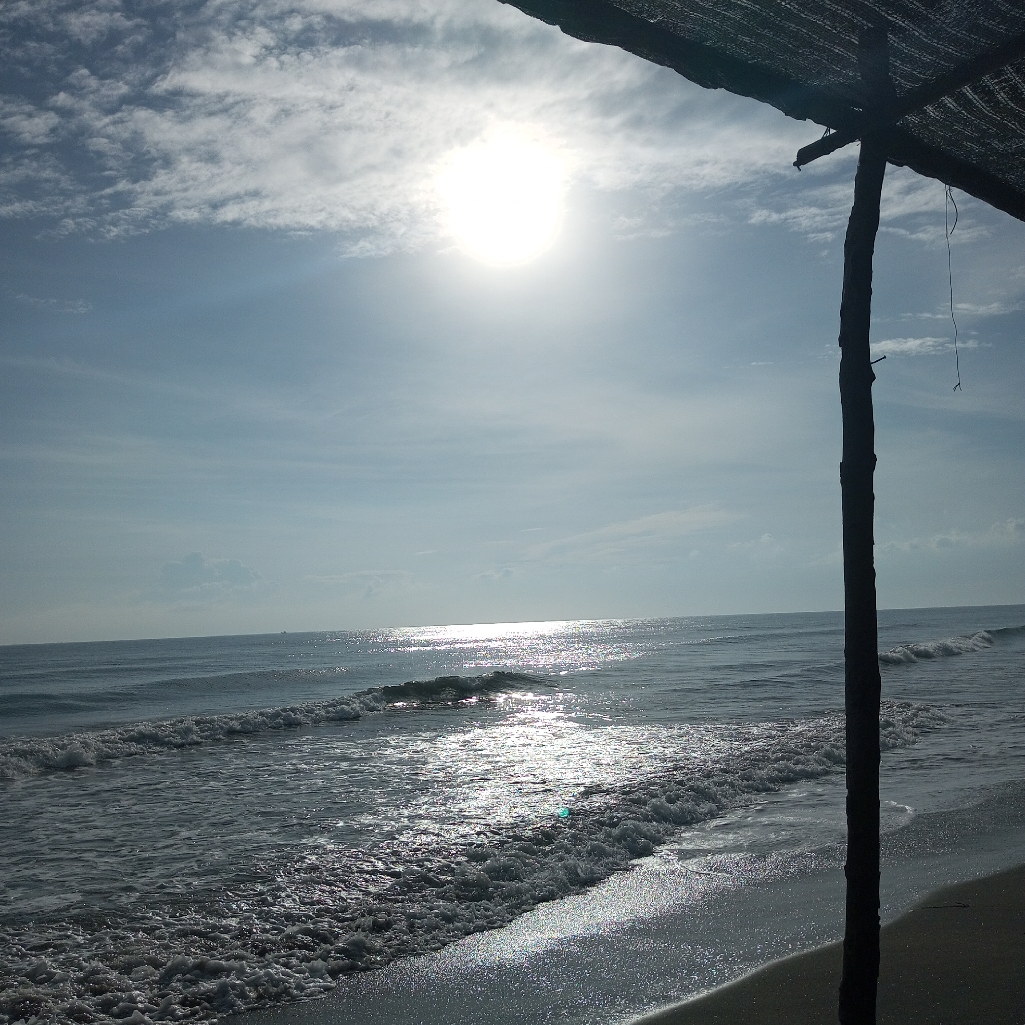 This media file captures a bright, sun-drenched beach scene, likely from Lhokseumawe, Indonesia. The time of day appears to be late morning or early afternoon, characterized by intense sunlight.
The scene features the ocean meeting a sandy shore under a clear, light blue sky dotted with scattered white clouds. The sun, a brilliant white orb, is prominently displayed high in the sky, casting a strong, shimmering path of light across the ocean's surface. Gentle waves break onto the shore, creating frothy white foam that sparkles brightly with the sun's reflection. The wet sand on the beach also glitters from the direct sunlight.
On the right side of the frame, a dark, weathered wooden pole stands vertically, partially obscuring the view of the beach and ocean. This pole is part of a rudimentary structure, supporting a dark, mesh-like roof, possibly a shade covering or a simple shelter. A thin string hangs from this structure near the pole. The image conveys a sense of natural beauty and tranquility. There are no people or animals visible, and no text is present.