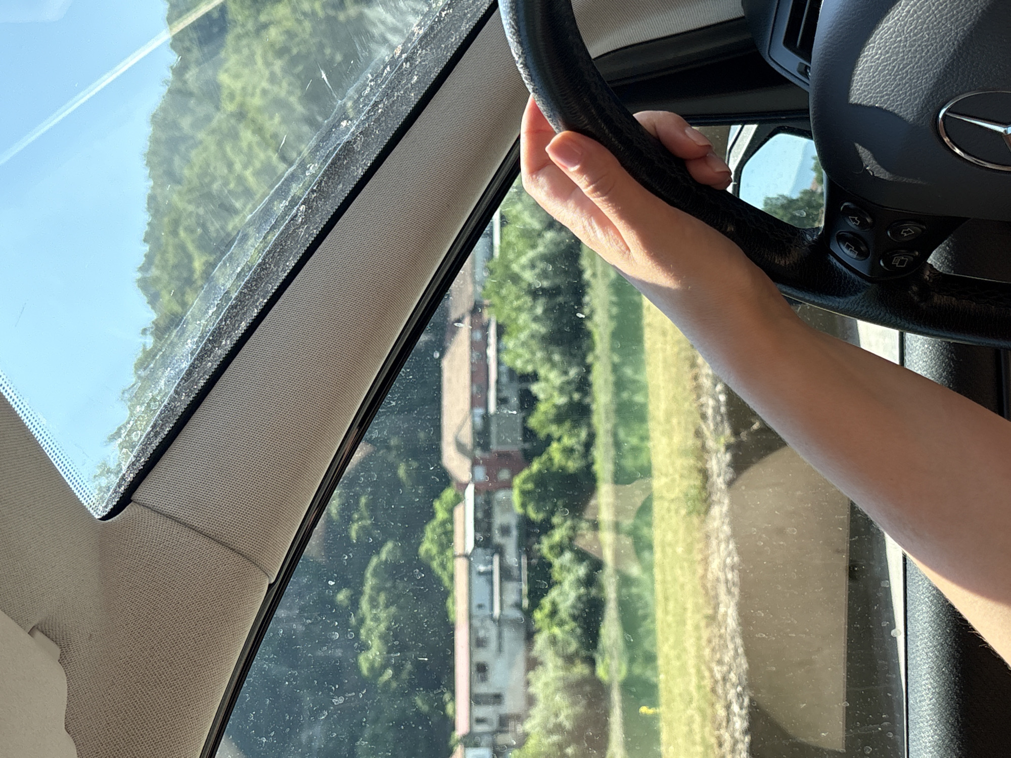 A first-person perspective shot from inside a car shows a person's hand on the steering wheel. Outside the vehicle, a body of water is visible, with a town situated on the opposite bank, nestled at the foot of a lush, green hillside under a clear blue sky. The town's buildings, featuring terracotta roofs, are clustered together. The scene appears to be along the Drina River, potentially in or near Zvornik, Bosnia and Herzegovina, given the mountainous terrain and the presence of the river. The windshield shows some dirt and water spots.