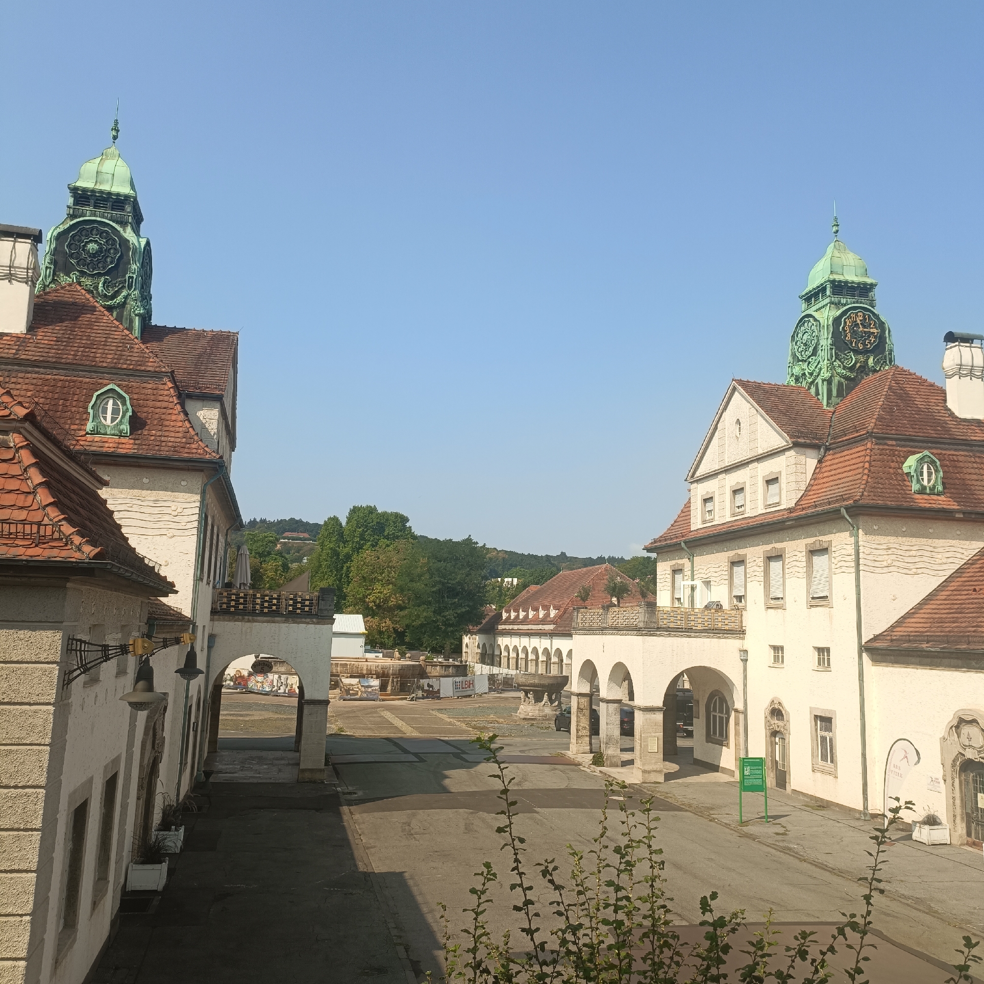 The image captures a sunlit scene in the city of Bad Nauheim, Germany, likely in the Kurpark area, showcasing the architectural grandeur of its historic spa buildings. The foreground features a paved courtyard with a low wall and a few scattered plants in the lower right and lower center. Two symmetrical, white-stucco buildings with red-tiled roofs dominate the sides of the frame. Each building is topped with a distinctive green-copper clock tower, one on the left and one on the right, indicating the passage of time. Archways connect various sections of the buildings, creating shaded walkways and revealing glimpses of the area beyond. The sky is a clear, bright blue, suggesting a warm, pleasant day. In the background, a row of more arched structures forms a colonnade, and beyond that, lush green trees and rolling hills are visible under the expansive sky. The architecture suggests a turn-of-the-century European spa or public building style, characterized by ornate details and prominent clock towers.