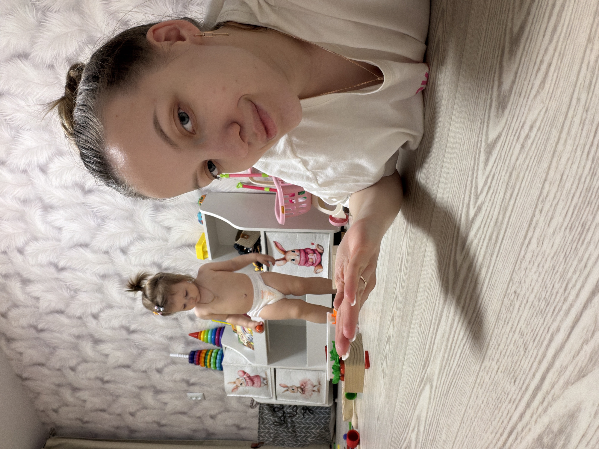 A woman and a young child are the subjects of this photograph, which appears to have been taken indoors. The woman is lying on a wooden floor, and her face is prominently displayed in the upper left corner of the image. She has fair skin, light eyes, and her hair is pulled back in a bun. She is wearing a white shirt and a gold necklace. The child, appearing to be a toddler, stands near a small, white toy cabinet on which the woman's hand is resting. The child is wearing a diaper and a necklace, and has their hair in a small ponytail with a flower clip. The cabinet is filled with various toys. The background features a white, textured wall. Based on the setting and the photographer's profile, it can be inferred that the location is likely in Reshetnikovo, Russia.
