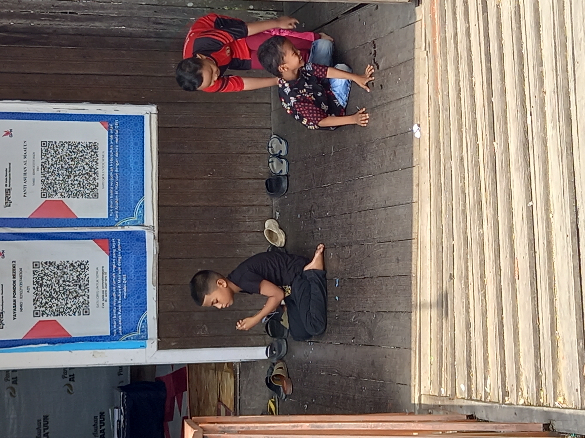 Three young boys are on a wooden porch, likely in Samarinda, Indonesia. The boy on the left, wearing a black t-shirt and black pants, is sitting cross-legged and appears to be praying or meditating. In the center, a boy in a patterned shirt and blue jeans is kneeling and reaching towards something on the wooden floor, possibly small insects or objects. The third boy, on the right, is standing and wearing a red and black striped shirt and pink pants, with his hand on the kneeling boy's back. Behind the boys, a wooden wall with two posters displaying QR codes and text in Indonesian is visible. Several pairs of sandals are scattered on the porch near the boys. The porch is elevated with a wooden railing on the left.