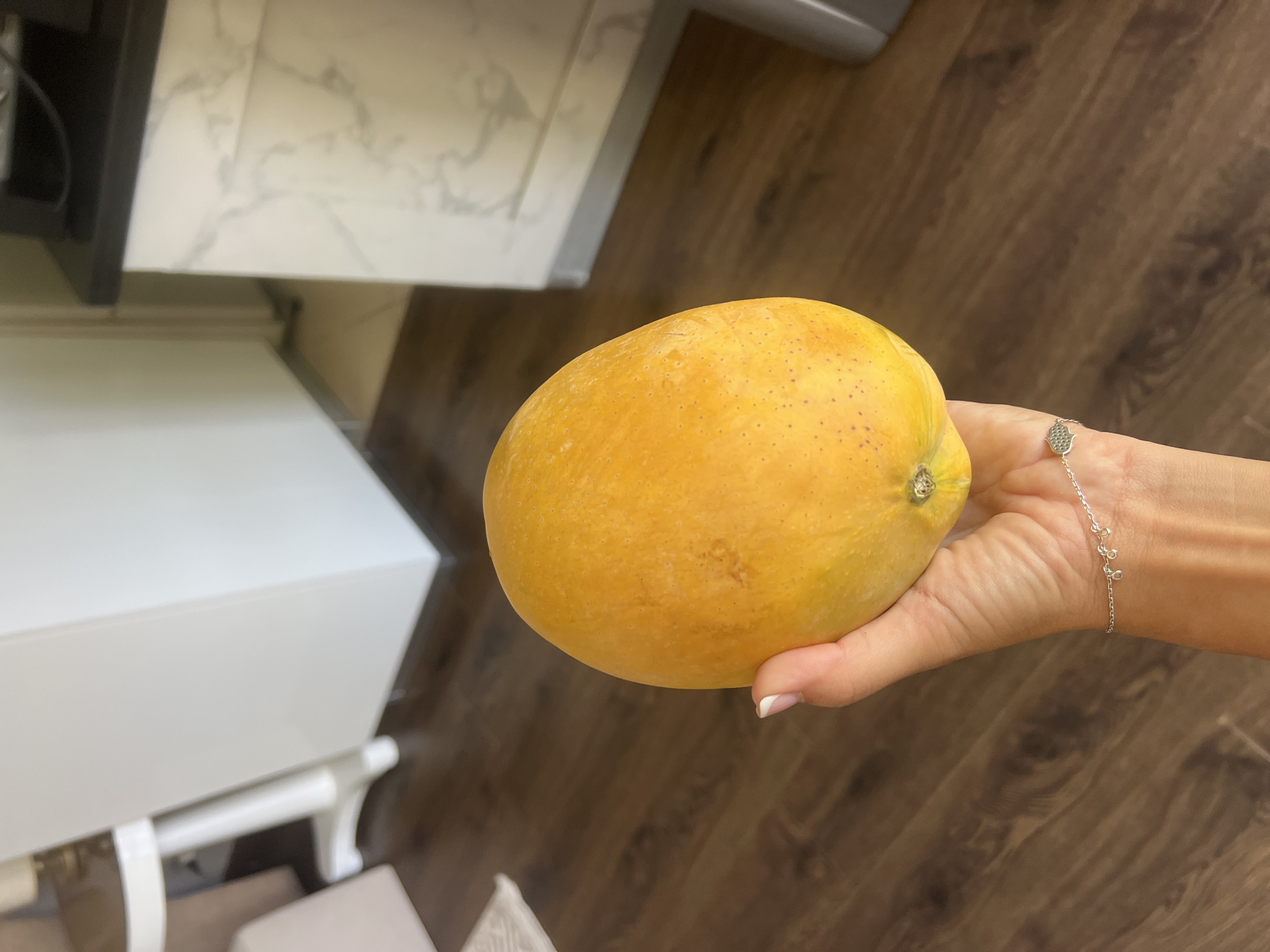 A close-up, eye-level shot shows a person's hand holding a ripe, yellow-orange mango. The mango is oval-shaped with a slightly irregular surface and small, dark speckles. The hand, with manicured fingernails, is positioned beneath the fruit. A delicate silver bracelet with small dangling charms is worn on the wrist. The background is blurred, revealing a dark wooden floor and hints of kitchen cabinetry with a marble-like pattern. The overall impression is one of a domestic setting. The image does not provide any information about the city of Sharm El Sheikh, Egypt.