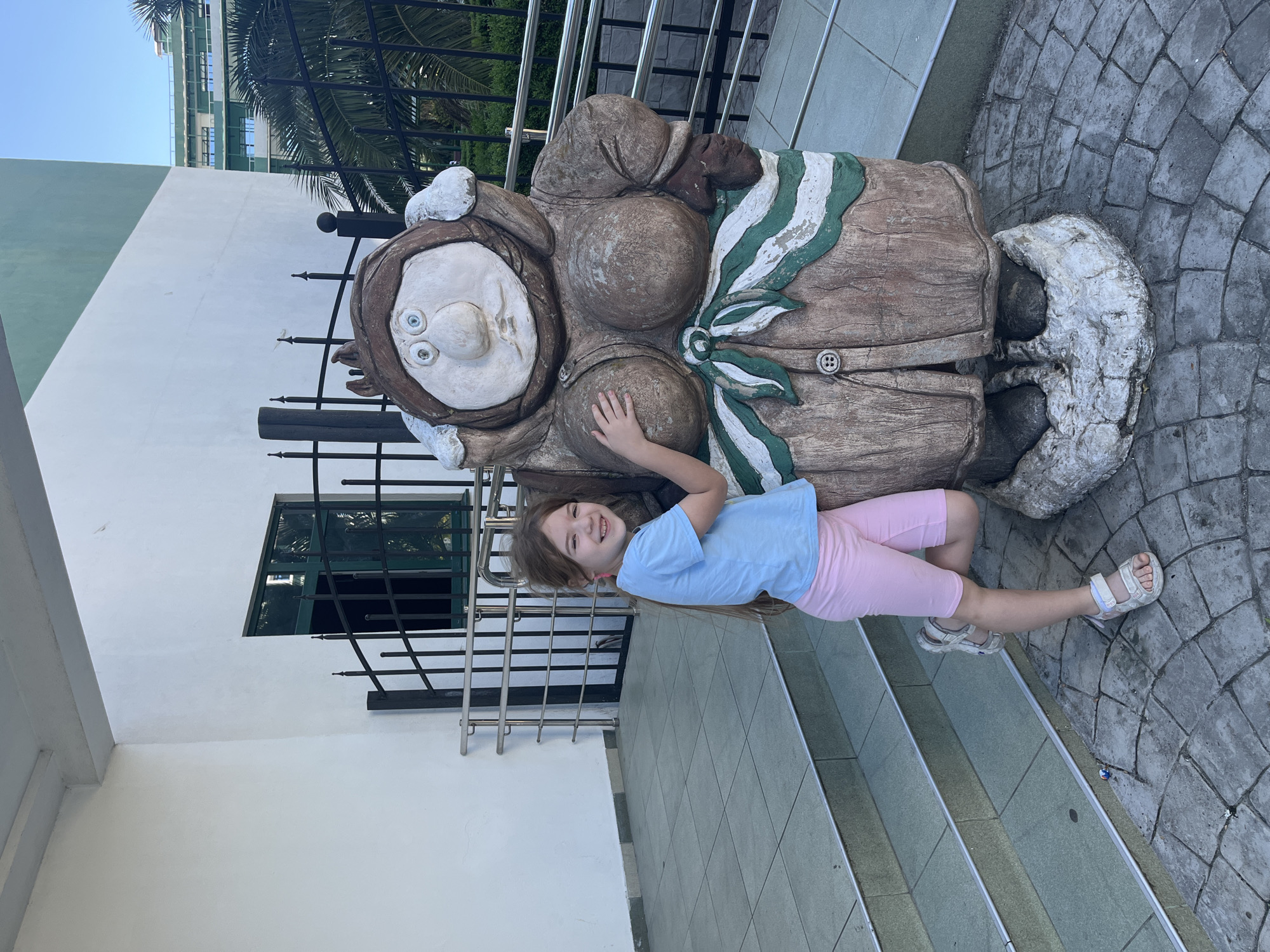 A young Caucasian girl with long brown hair, wearing a light blue t-shirt and pink shorts, stands with her left leg bent and resting against a large, weathered statue of a female figure in Sochi, Russia. The statue appears to be made of concrete or stone and depicts a plump woman wearing a brown coat with a green and white sash. The girl is smiling at the camera and has her right hand resting on the statue's chest. The statue is positioned on a cobblestone area, with stairs leading up to a building with white walls and a green-trimmed upper section in the background. A metal railing and fence are also visible. The sky is clear and blue.