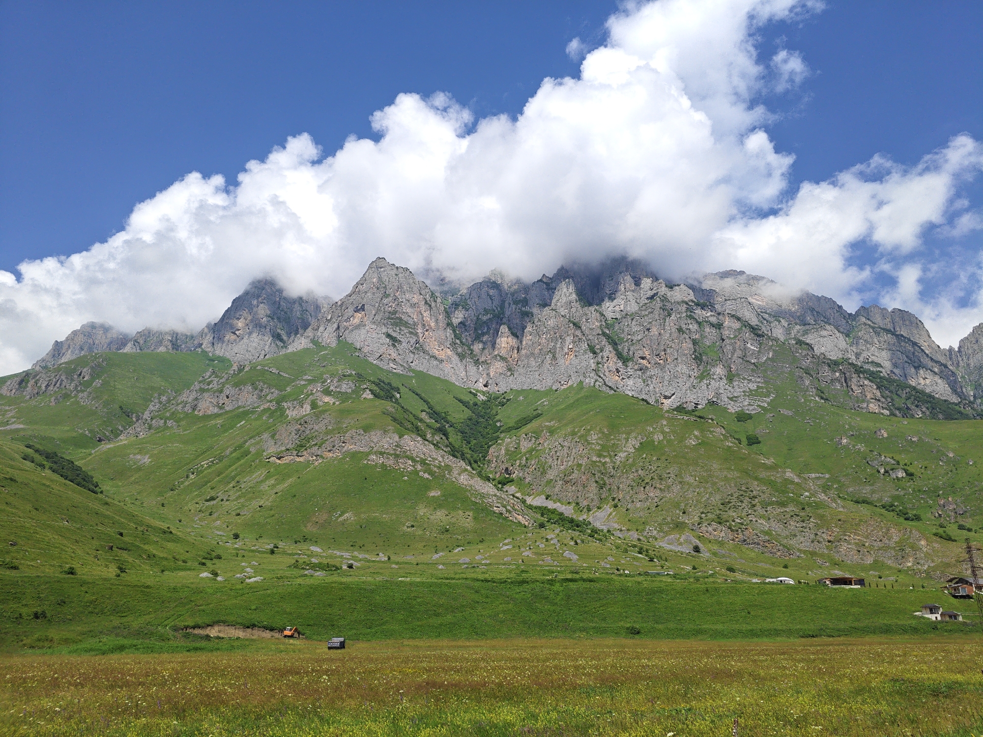 The image depicts a vast, mountainous landscape under a partly cloudy blue sky. In the foreground, a meadow covered with wildflowers stretches across the frame, with patches of taller green grass. In the mid-ground, a gently sloping green hill is visible, dotted with scattered rocks. Further back, the imposing, rugged mountains rise dramatically, their rocky faces partly obscured by a thick blanket of white clouds. The slopes of the mountains are covered in green vegetation, with darker patches indicating ravines or denser foliage.
On the green hills in the mid-ground, several structures are visible. On the left, an orange excavator is parked on the slope. To its right, a dark-colored vehicle is present. Towards the right side of the mid-ground, a cluster of buildings, appearing to be houses, are situated on the hillside, with a tall electrical pylon nearby.
The scene is one of natural grandeur, with the mountains forming a powerful backdrop to the cultivated and natural elements in the foreground and mid-ground. The presence of human infrastructure, such as the buildings and the excavator, suggests this area is inhabited or being utilized. The location is identified as being in the vicinity of Dargavs, Russia, a region known for its scenic mountainous terrain and historical significance.
