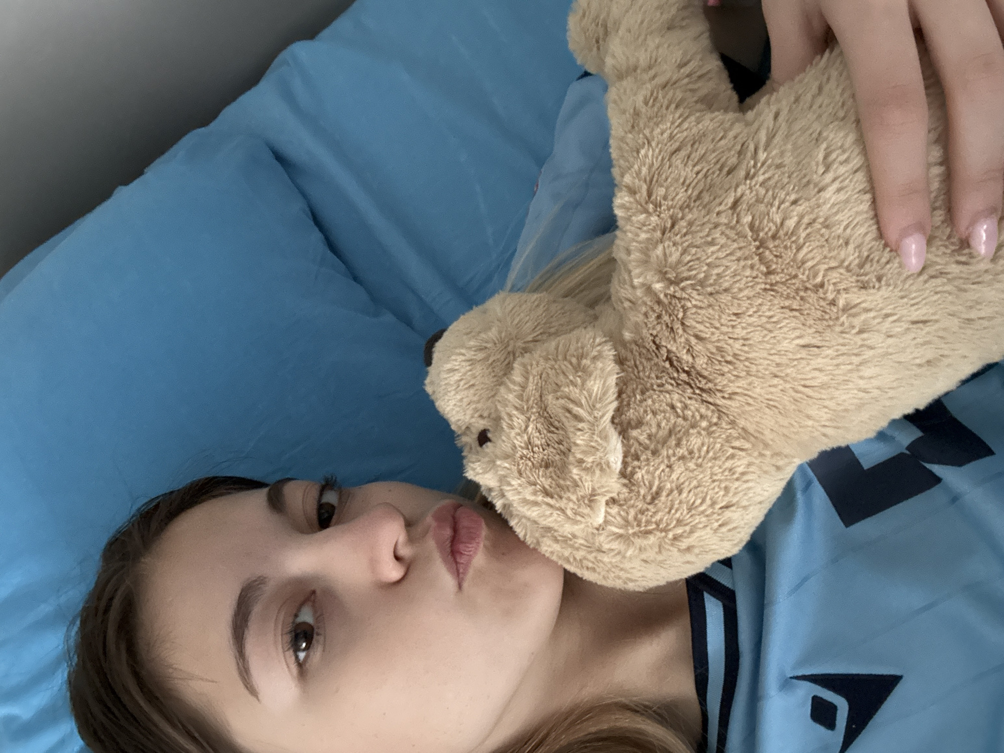 A fair-skinned young woman with brown hair is lying on a blue bed or possibly a couch, holding a tan stuffed animal dog. Her eyes are brown, and she's wearing minimal makeup. She has a neutral expression. She is wearing a blue shirt with black details. The background consists of blue bedding. The location is not discernible from the image and thus cannot be identified as being in Sofia, Bulgaria.
