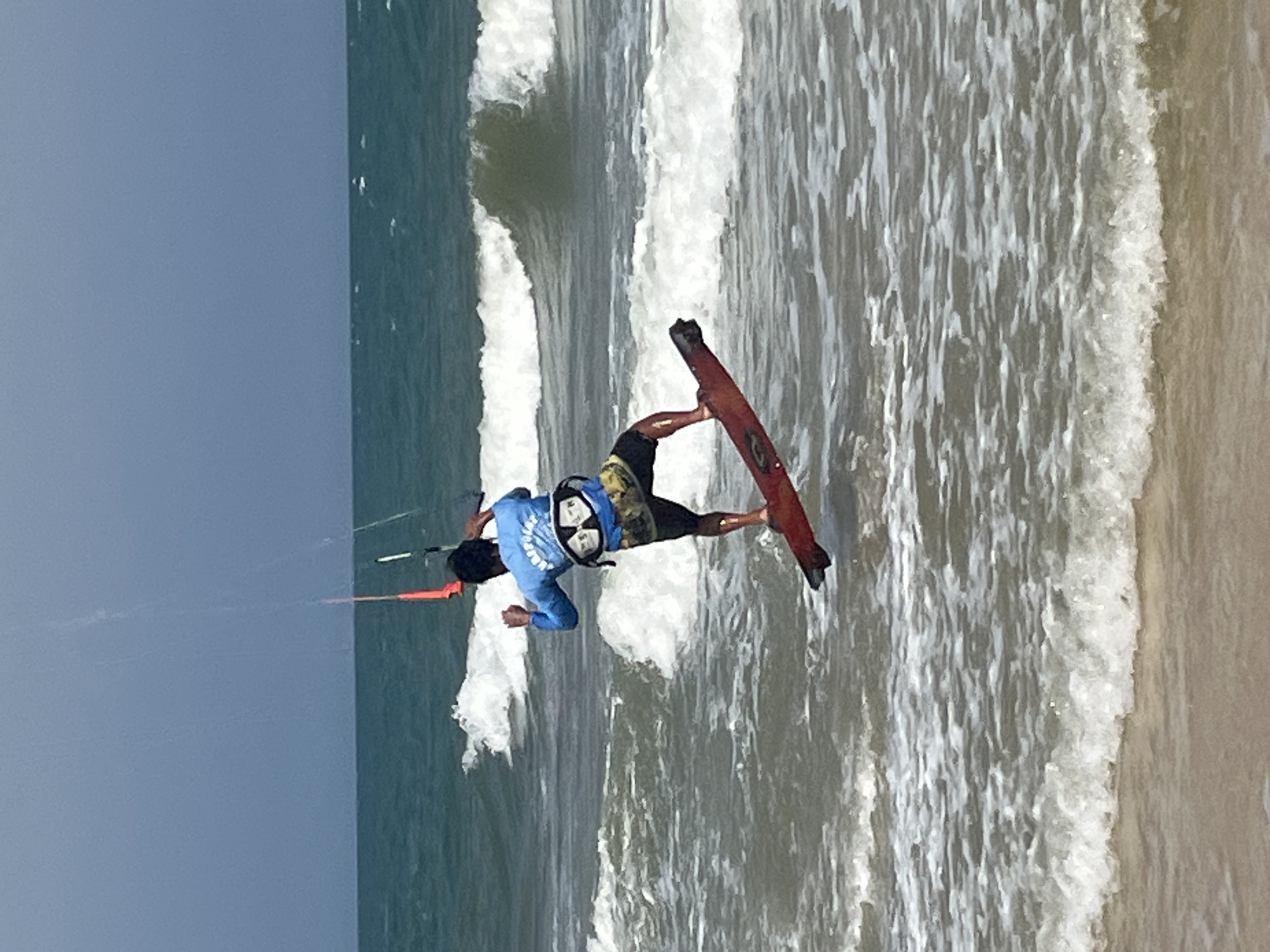 A person is kitesurfing in the ocean off the coast of Phu Thuy Ward, Vietnam. The kitesurfer is in mid-air, having launched off a wave, with their kite tethered above them. They are wearing a blue long-sleeved shirt, a life vest, and patterned shorts. The kitesurfing board is red and is positioned beneath their feet. The ocean is blue-green with white waves crashing. The sky is overcast and a pale blue. The shore is visible in the foreground, appearing as a sandy beach.