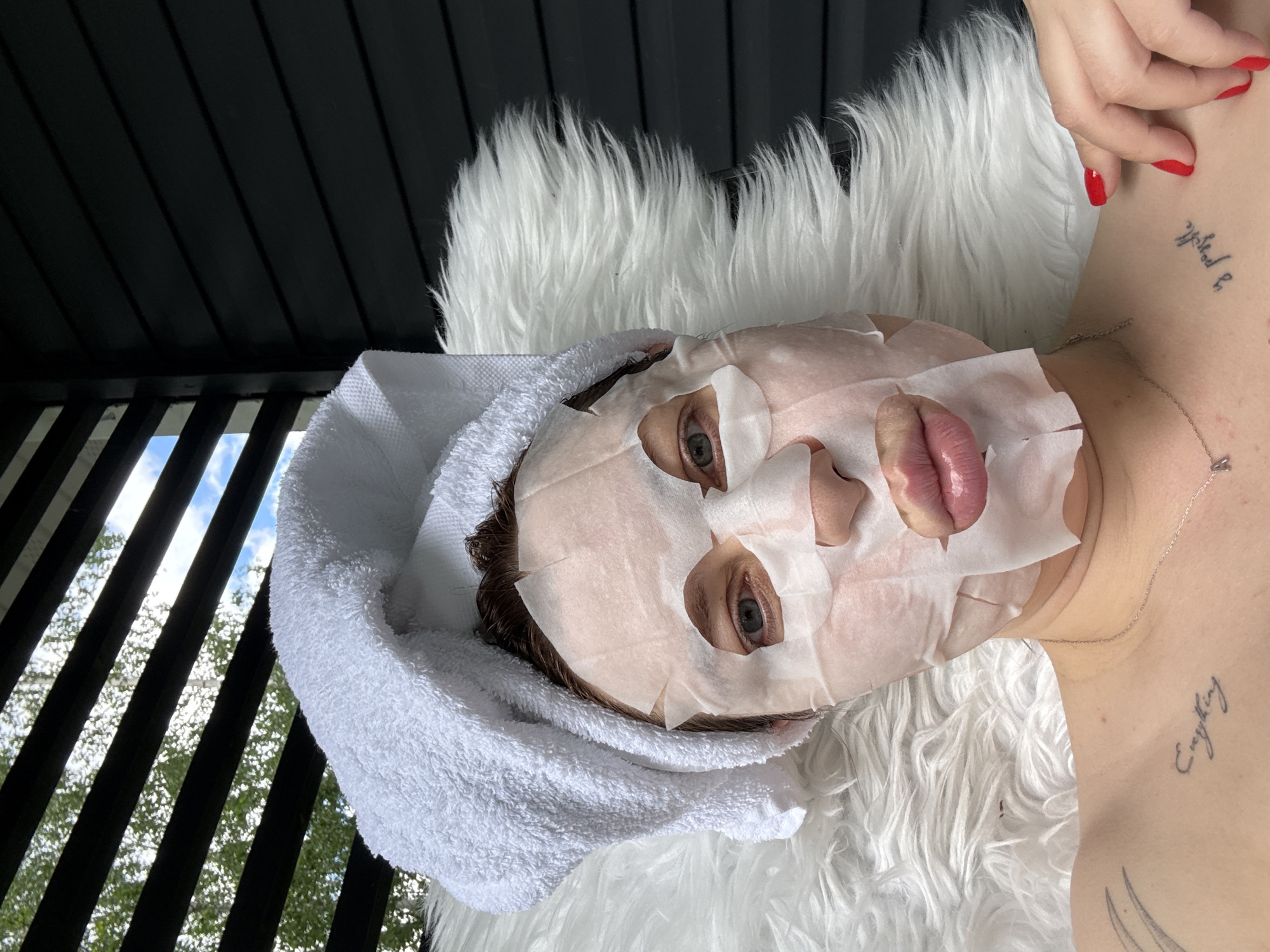 A person wearing a facial sheet mask and a towel turban is lying on a white fur-like surface. The person has visible tattoos on their neck and chest. The background includes a slatted structure, possibly a balcony or veranda, with glimpses of trees and a cloudy sky. The photo appears to be taken indoors. The location in the city of Buzhaninovo, Russia is not deducible from the image.