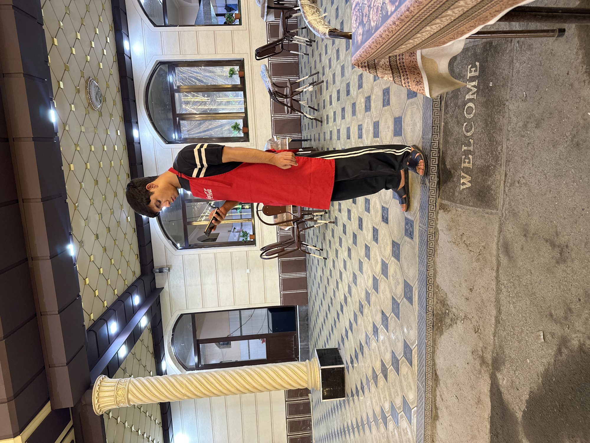 A young male wearing a red apron and black pants with white stripes is standing on a patterned tile floor, looking at a smartphone. He is in an outdoor seating area of what appears to be a restaurant or cafe. The area is covered by a decorative ceiling with a geometric pattern and recessed lighting. There are pillars, tables, and chairs visible in the background, along with windows and doorways. A welcome mat is present on the ground in front of the seated figure. The lighting suggests it is evening or a dimly lit indoor space.