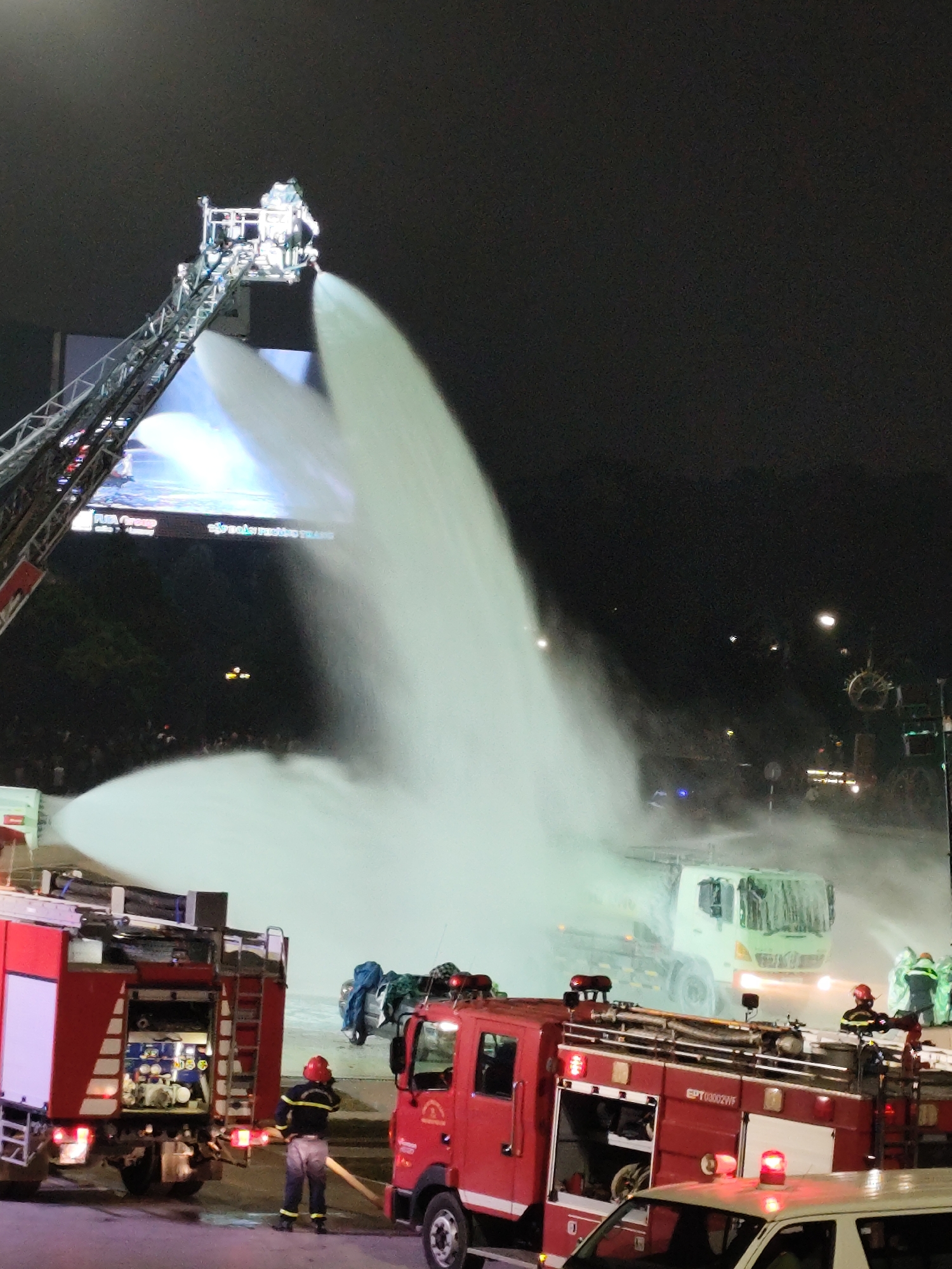 The image depicts a nighttime scene in Da Lat, Vietnam, likely a street or open area, where a large-scale water spray is occurring. A fire truck with a high-reach ladder is prominently positioned, with a powerful stream of white water or foam being directed from its elevated platform. Below the ladder, several other fire trucks, distinguished by their red color, are visible, along with some vehicles and what appears to be a crowd of onlookers or emergency personnel in the background, partially obscured by the water spray. Several individuals in firefighter gear are present, some operating equipment and others observing the activity. The overall impression is one of an emergency response or a public demonstration involving a significant water display, set against a dark, urban night sky.