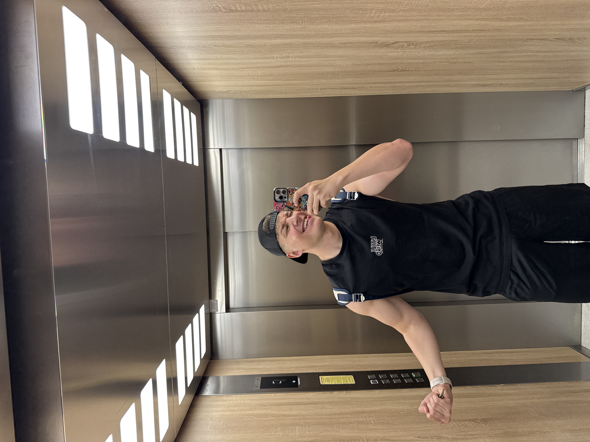 A young adult male is captured in a selfie within a modern elevator, displaying a broad, joyful smile. He is dressed in a black t-shirt with "MELKO TRIBE" printed on the chest, black shorts or athletic pants, and a black baseball cap worn backward. On his left wrist, he wears a white wristband or watch. He holds a smartphone with a multi-colored case in his raised right hand, positioned to take the picture. His left hand is slightly clenched, holding a set of keys.
The setting is an elevator interior, characterized by sleek, metallic walls that appear to be polished stainless steel, and a light-colored wooden ceiling and floor. One side wall features a series of bright, vertical rectangular lights providing illumination. On the right side of the elevator, a control panel is visible, integrated into the wooden surface. It includes several square buttons, a small display showing "L J" (possibly indicating the floor or direction), and a yellow cautionary sticker with unreadable text. The man's reflection is faintly visible in the polished metal elevator doors behind him. The scene suggests a casual moment, possibly in Plovdiv, Bulgaria, where the man is playfully taking a selfie.
