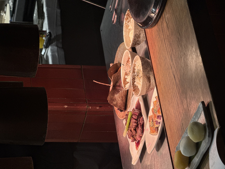 The scene depicts an interior view of a restaurant in Paris, France, focused on a food service area. In the foreground, a light-colored wooden counter extends horizontally. On this counter, a rectangular grey plate holds three spherical mochi-like desserts: one yellow, one white, and one light green. A portion of a white paper document is visible beneath the plate.
Behind the wooden counter, a metallic or stainless steel surface displays several food items under warm, localized illumination from two dark, conical overhead lamps. A white rectangular plate contains slices of seared or grilled meat, possibly beef, resting in a red sauce and garnished with a single green stalk. Below this, another white rectangular plate holds multiple pieces of cooked fish, possibly salmon, seasoned with dark flakes. Behind these plates, a larger round wooden plate presents an item wrapped in a large, dried brown leaf, secured by a small stick. Three small, textured ceramic bowls, varying in color from light grey to beige, contain white rice topped with dark seasoning. To the far right on the metallic surface, a portion of a round, dark, metallic object with a handle is visible, likely a food warmer or cooking utensil.
In the mid-background, a reddish-brown tiled column stands vertically, exhibiting some surface wear. Further into the background, a table draped with a white tablecloth is partially visible, with indistinct objects placed upon it. The overall lighting is dim, creating a focused, warm glow on the food items against darker surroundings. No individuals are present in the frame.