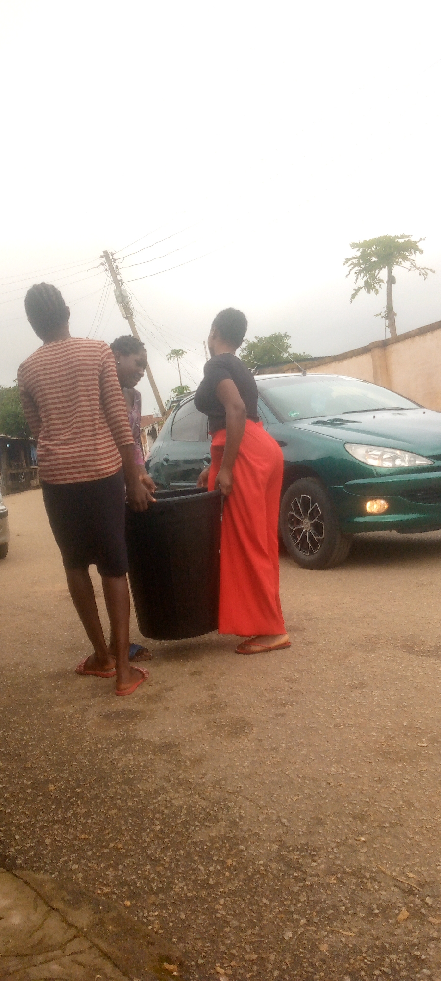 Three young women are standing on an asphalt road, one of whom is holding a large black bucket. The woman on the right wears a black top and long, flowing red pants, while the woman on the left wears a striped long-sleeved shirt and a dark skirt. The third woman, partially obscured, stands between them. A dark green car is parked behind the women. In the background, there are power lines, a pole, a fence, and some trees, including a tall, slender tree with a few branches at its top. The sky is overcast and light-colored. The scene appears to be in an outdoor, possibly urban or semi-urban, setting.