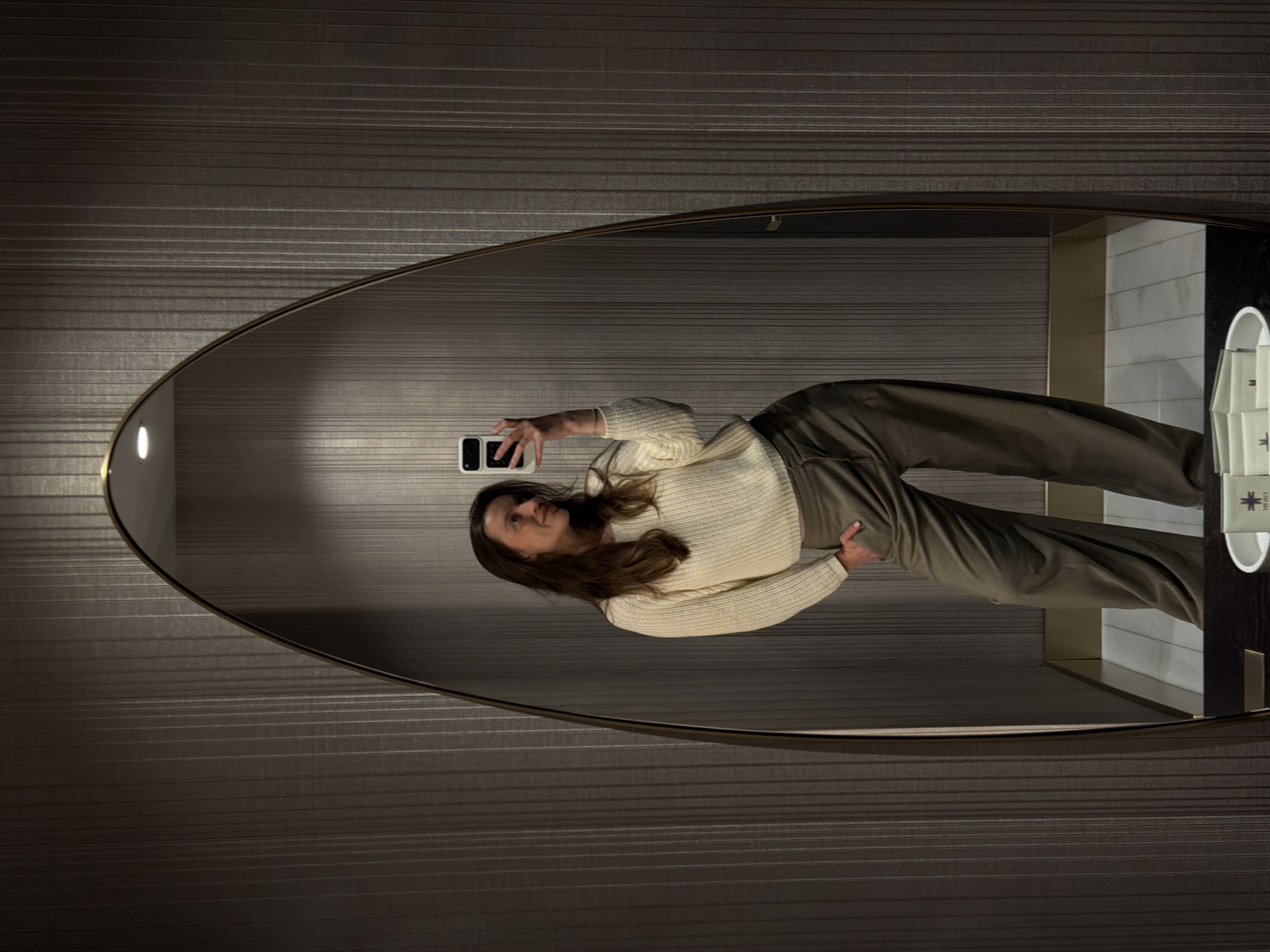A person is reflected in an oval mirror. The person is holding a black mobile phone and wearing a white sweater and olive-green pants. The background is a dark, textured wall with horizontal lines. The mirror is located in a bathroom in Budva, Montenegro.