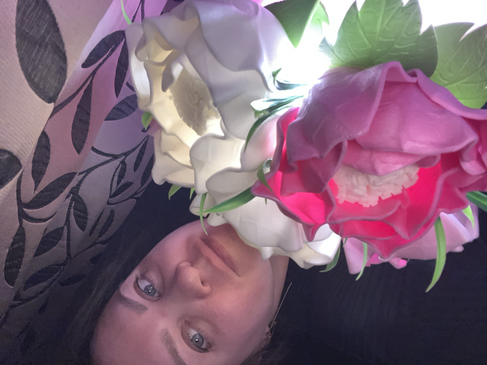 A portrait-style image features a woman's face positioned beneath two large, prominent artificial flowers. The woman has light skin, blue-grey eyes, and dark hair, looking directly at the camera with a neutral to slightly pensive expression. Her face is illuminated, highlighting her features. Above her, to the right, are two decorative artificial flowers: one a bright white/cream and the other a vibrant pink, both accented with green leaves. These flowers appear to be internally lit or backlit, casting a soft glow. The background behind the flowers and the upper part of the woman's head is dark, providing a strong contrast to the illuminated subjects. To the left of the woman, a patterned fabric with dark, leaf-like motifs on a lighter, possibly cream or light pinkish, background is visible. The scene suggests an indoor setting, possibly a self-portrait taken with decorative lighting. No text is visible.