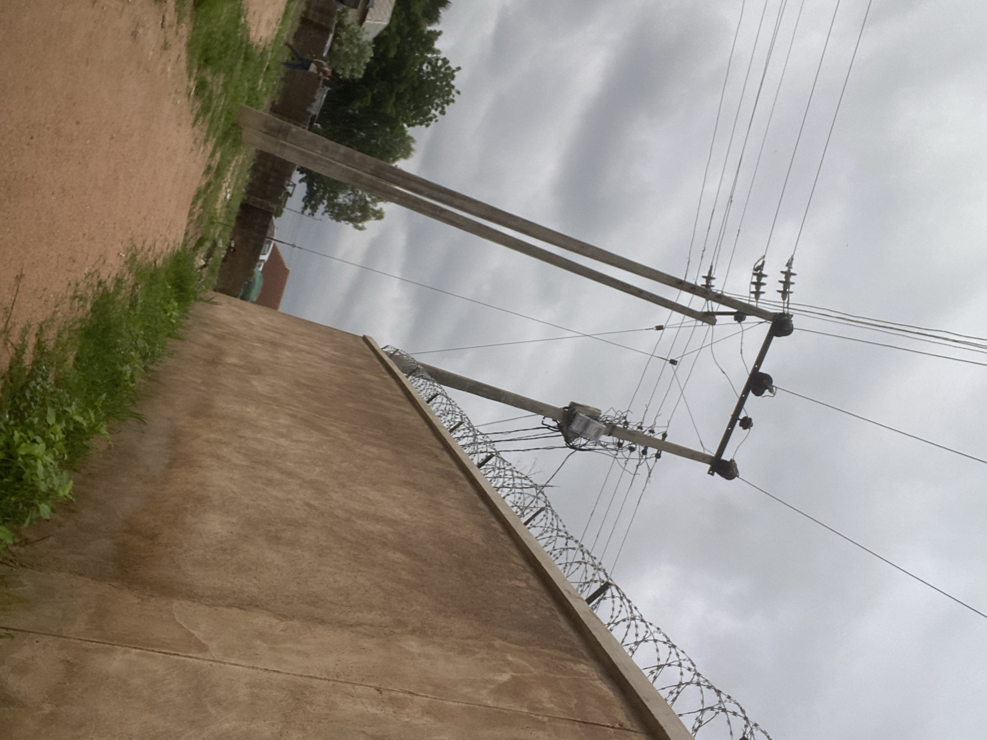 The image, taken in Gbaza, Nigeria, depicts an outdoor scene with a focus on a section of a beige wall topped with barbed wire. The wall appears to be part of a boundary or enclosure. Above the wall, a utility pole with electrical wires is prominently visible, stretching across the frame against a cloudy sky. The sky suggests it might be a grey or overcast day, possibly late afternoon or evening given the diffused light.
To the left of the wall, a dirt path runs alongside it, with patches of green grass growing along the edge. In the background, glimpses of trees and structures can be seen, hinting at a semi-urban or rural setting. The electrical infrastructure, including the pole, insulators, and numerous wires, suggests the presence of electricity supply to the area. The barbed wire on top of the wall implies a need for security or restricted access. The overall impression is one of a functional, everyday environment with visible signs of infrastructure and security measures. No people are clearly visible, and there is no discernible text in the image.