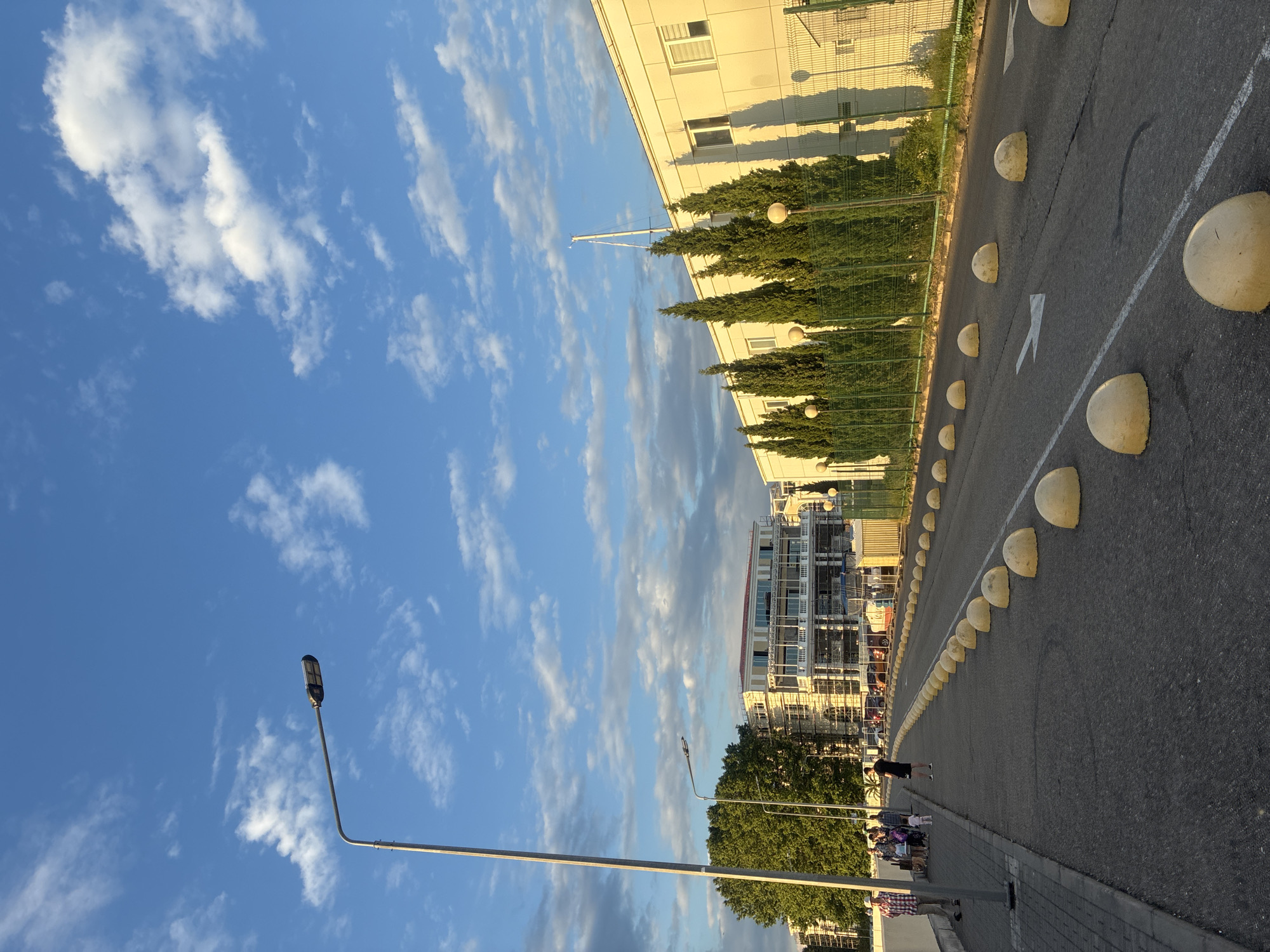 The image captures a street scene in Sochi, Russia, under a bright blue sky dotted with wispy white clouds, suggesting it's daytime, likely late afternoon given the warm light. The perspective is from a low angle, looking up and across a paved road.
Dominating the right side of the frame is a road marked with white lines and a series of yellow, half-sphere bollards. A few people are visible walking along the sidewalk next to the road. In the background, a modern building with glass windows and balconies stands out, alongside a line of tall, dark green trees. Further along the road, another building, seemingly under construction or renovation with scaffolding, is visible. A street lamp with a modern fixture stands prominently in the foreground, its pole extending upwards towards the sky. The overall atmosphere appears calm and casual, typical of a public outdoor space.