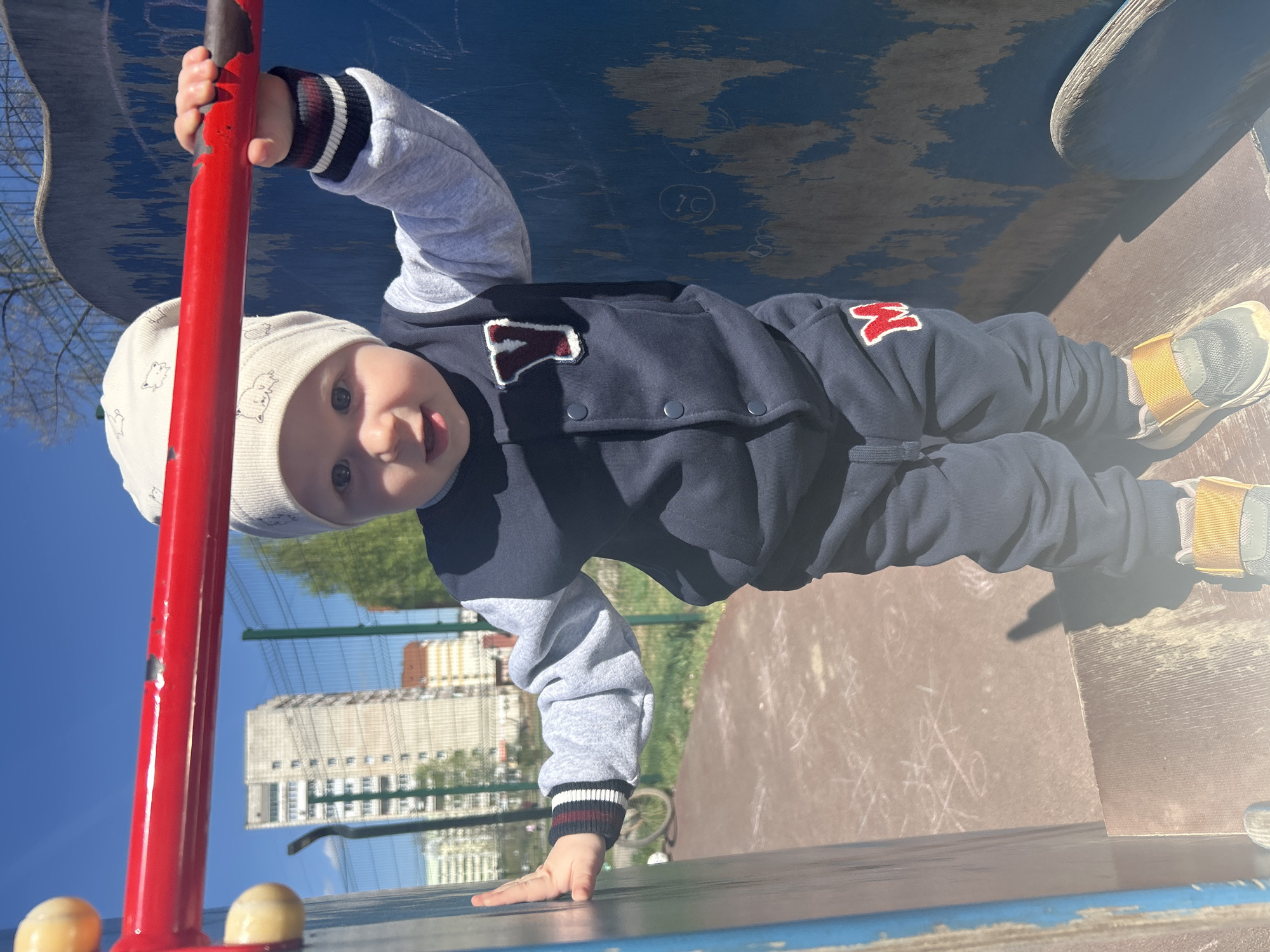 A baby is at a playground in the city of Chaikovsky, Russia. The baby is wearing a white hat, a dark blue jacket and pants with a grey long-sleeved shirt underneath, and grey shoes with yellow straps. The baby is holding onto a red metal bar on a playground structure. The playground surface is brown and blue, and there is a building in the background. The sky is blue. The location appears to be outdoors on a sunny day.