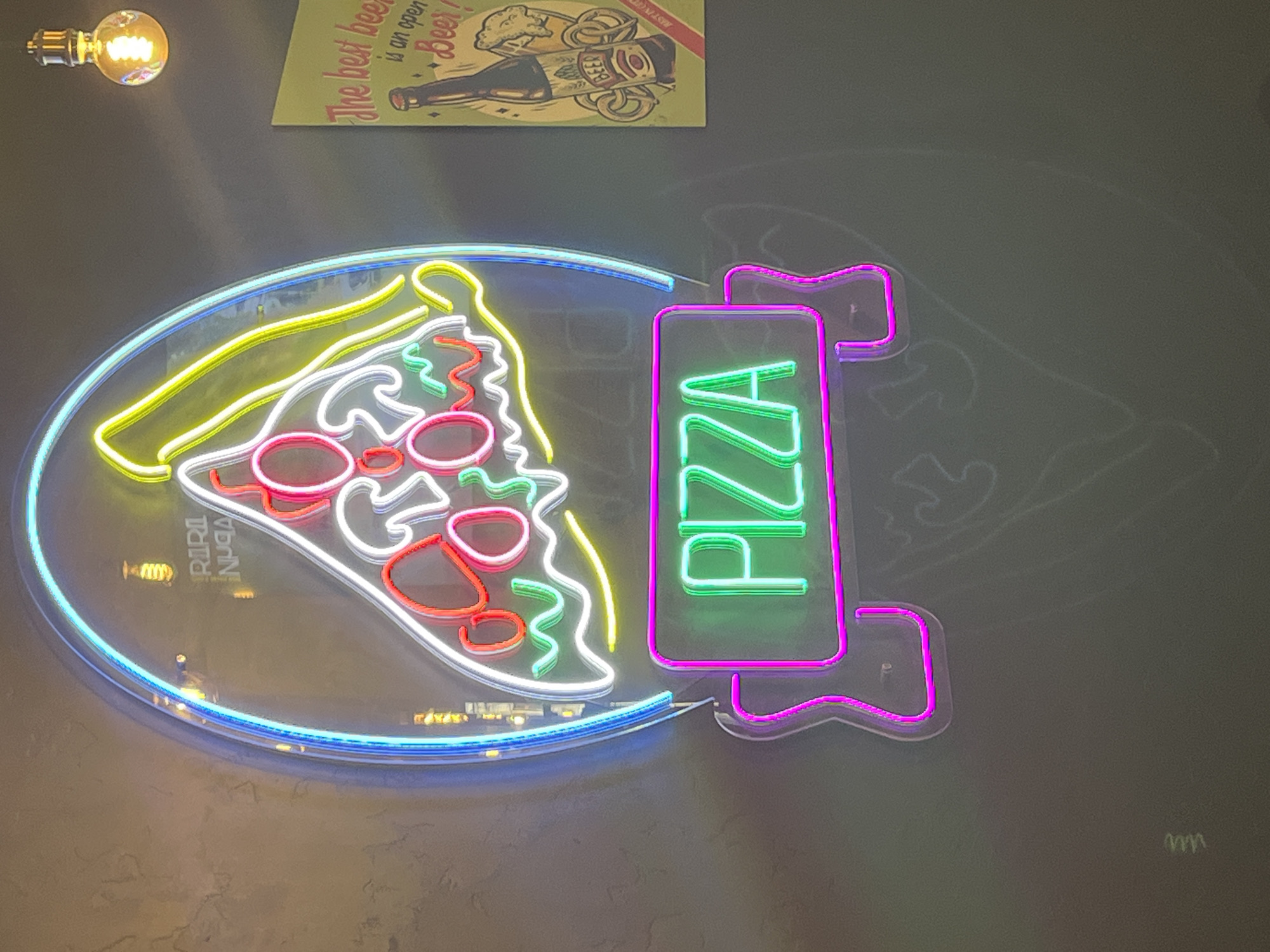 The image shows a wall decorated with neon signs, likely inside a restaurant or bar. The main sign is a stylized depiction of a pizza slice and the word "PIZZA" in a vertical arrangement. The pizza slice is outlined in blue neon, with yellow accents for the crust and white, red, and green to represent the toppings. "PIZZA" is rendered in green neon, outlined in pink. In the upper left corner is a light bulb, and above the pizza sign is a picture with the words "The best beer is an open Beer!". The photo was taken in Izhevsk, Russia.