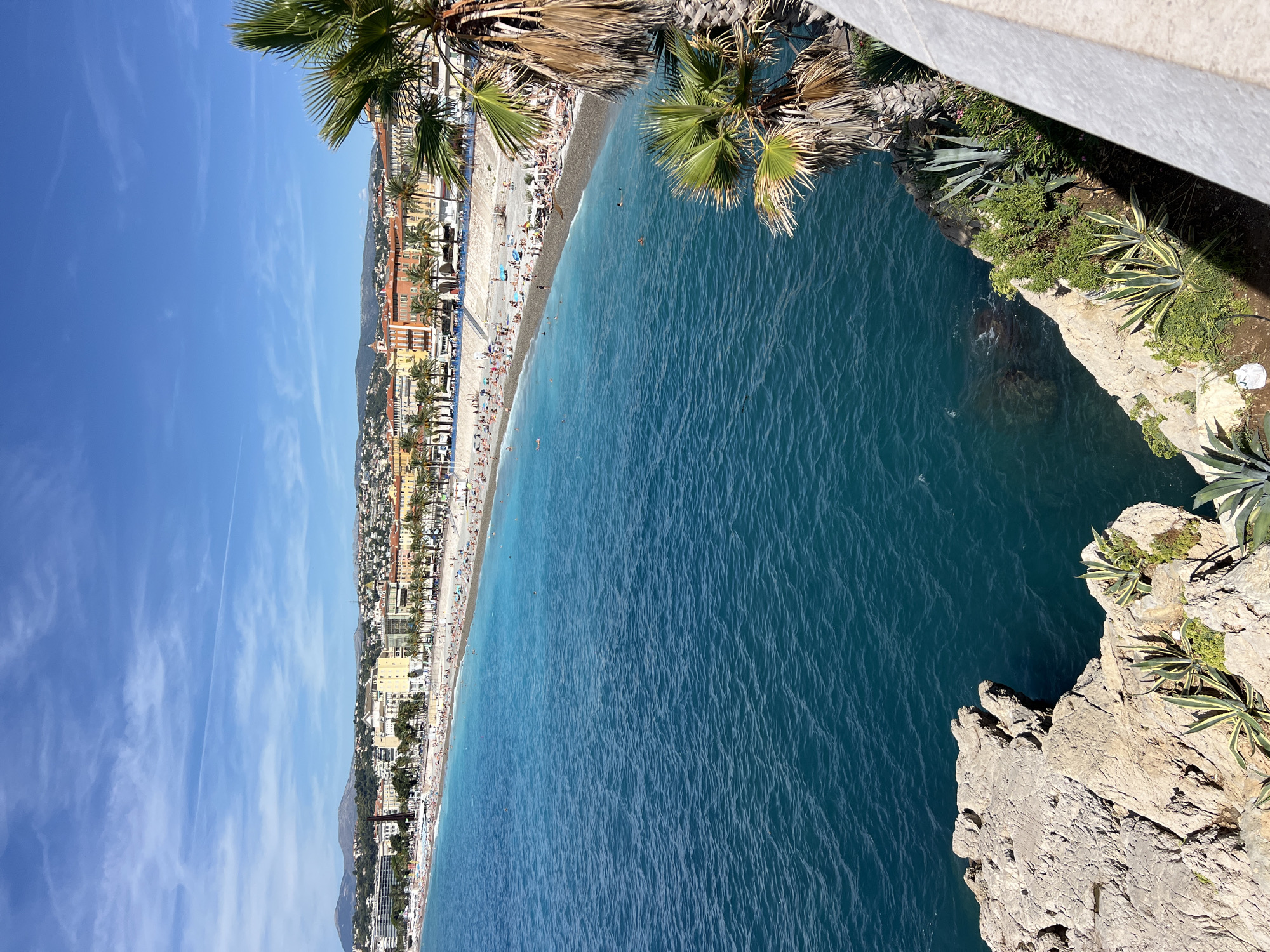 This is a high-angle, full-shot photograph of the coastline in Nice, France. The foreground features a rocky outcropping with sparse vegetation, including agave plants. To the right, a portion of a white railing is visible, casting a shadow. The middle ground displays a vibrant turquoise sea with gentle ripples, and a pebbly beach dotted with numerous sunbathers, umbrellas, and beach chairs. Palm trees line the edge of the beach, separating it from the city buildings. The background consists of a densely populated cityscape with colorful buildings, many featuring terracotta roofs, stretching along the coast. Mountains are visible in the distance, under a bright blue sky with scattered wispy clouds. The overall scene depicts a sunny day at a popular seaside resort.