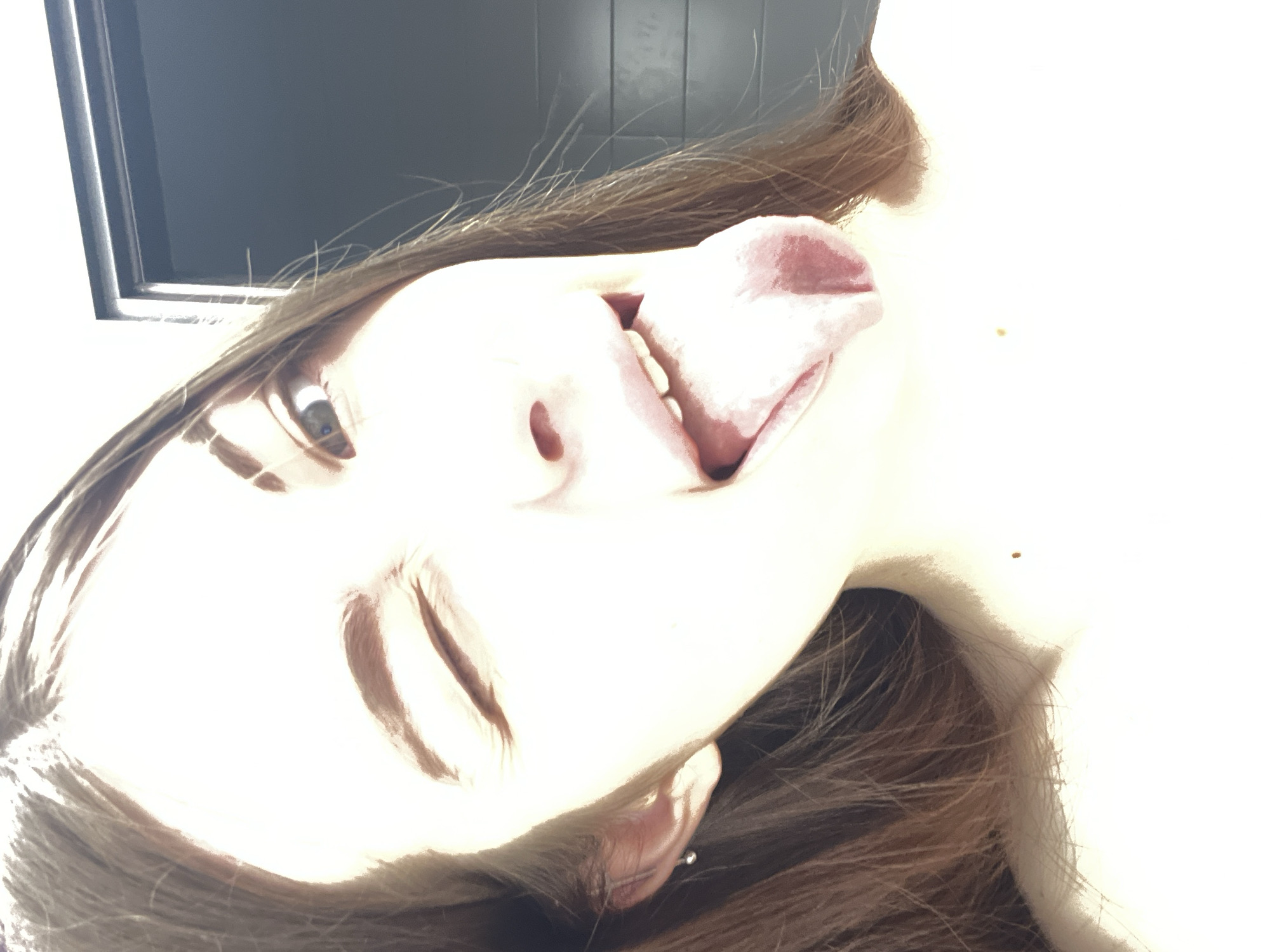 A close-up, low-angle shot depicts a person with brown hair and fair skin sticking their tongue out. The person's face is brightly lit, and they have one visible eye. The background is partially obscured by a window frame in the upper left corner. There is no visual information to discern that this image was taken in the city of Saint Petersburg, Russia.