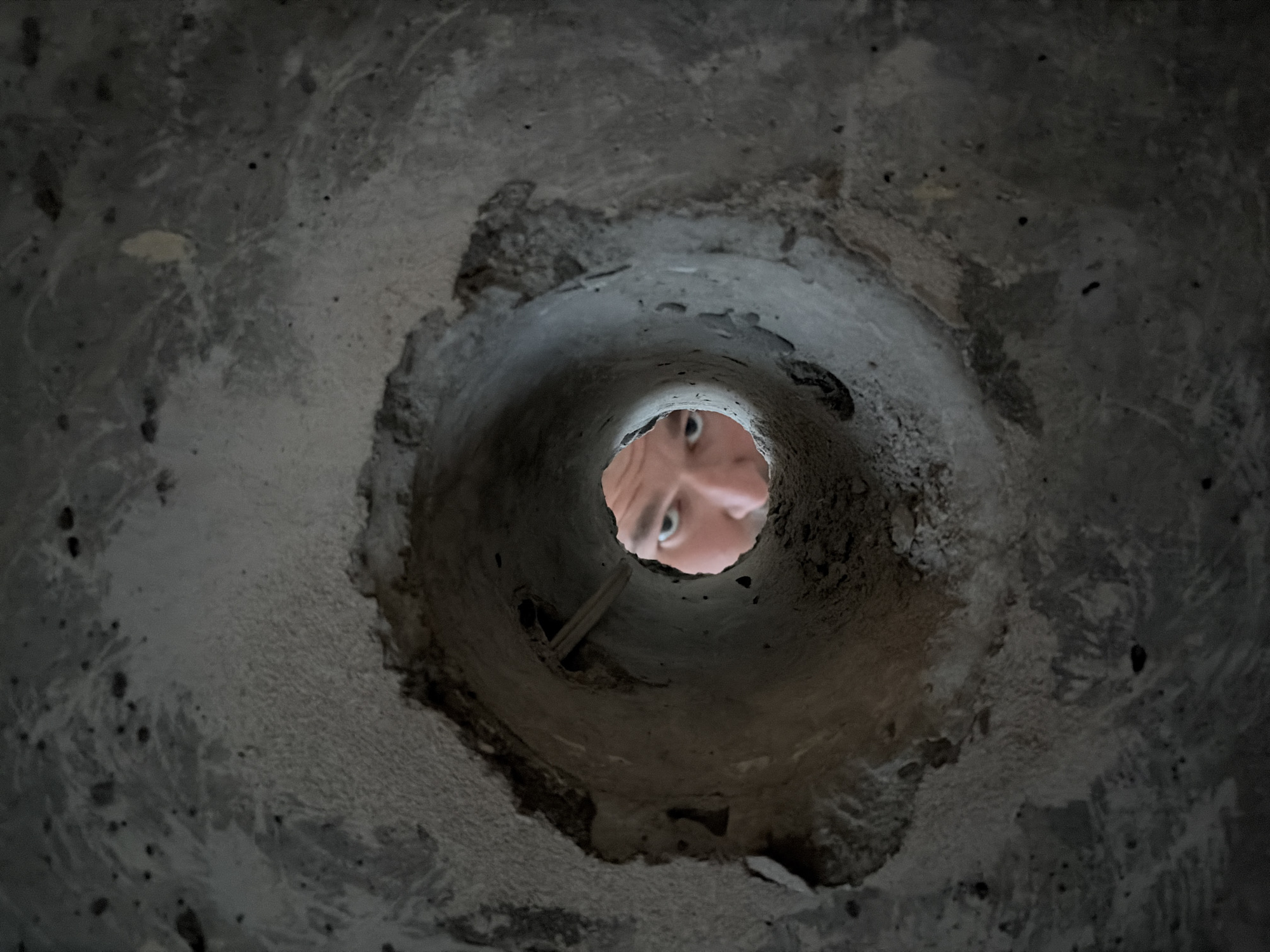The image is a close-up, low-angle shot looking into a roughly circular hole in a concrete-like surface. The interior of the hole is dark and appears textured. A person's face, specifically the eyes, is visible at the far end of the hole. The hole is irregularly shaped and seems to be made from a rough or broken material. There is no indication from the image that the location is Izhevsk, Russia.
