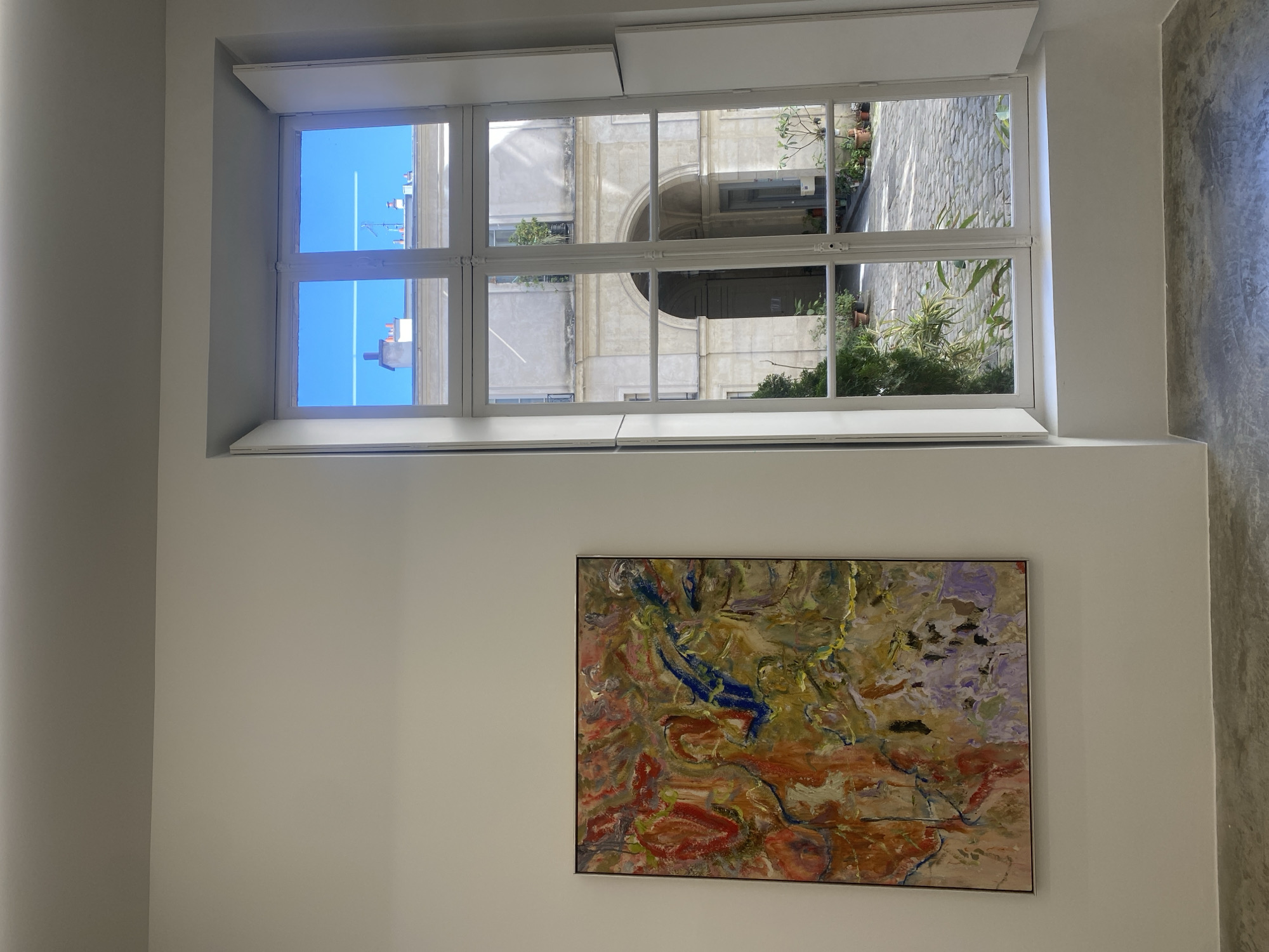 A framed painting hangs on a white wall beneath a partially opened window with white trim. The window is divided into panes, with the top section open and folded outwards. Through the window, an exterior scene of a Parisian courtyard is visible. The courtyard features a building with an arched entrance, along with greenery and a cobblestone-style ground. The sky is visible in some window panes. The painting below the window features a textured, abstract design with shades of red, yellow, blue, and lavender. To the right of the painting, a concrete wall is visible.