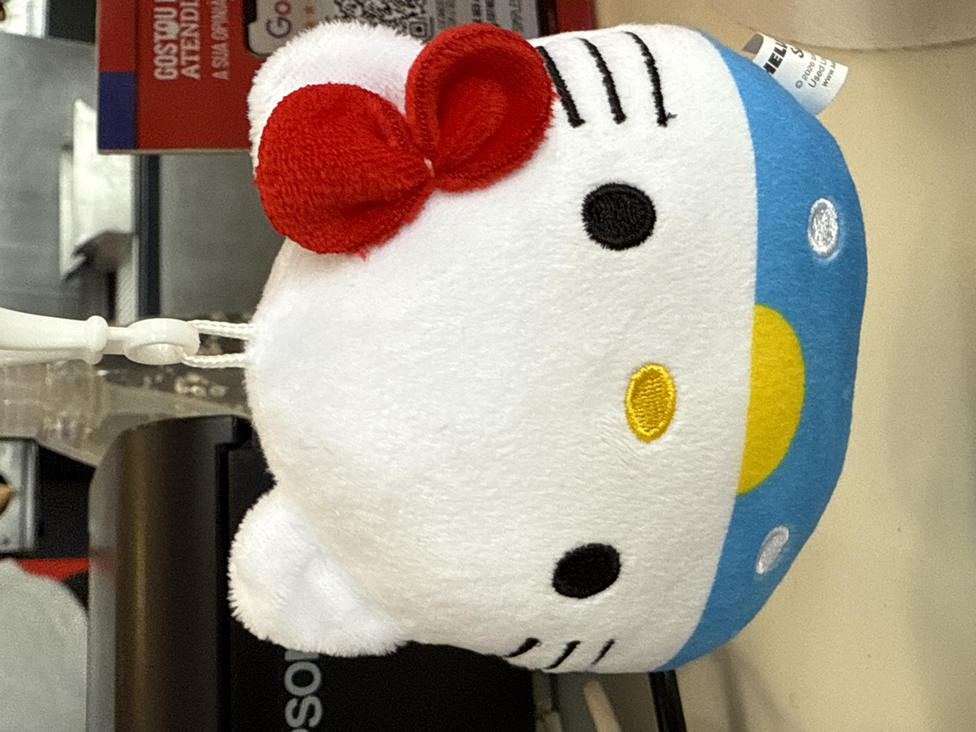 A close-up, eye-level shot shows a white plush Hello Kitty keychain with a red bow. The plush toy is hanging from a white clip. The Hello Kitty's face has black eyes, a yellow oval nose, and three black whiskers on each side. Its lower half is blue with a yellow smile and a small, white circular detail. In the background, a black printer is visible on the left, and a red sign with white text is on the right. A white paper tag is attached to the bottom right of the plush toy. The lighting is bright and natural. The scene is indoors, likely in a commercial setting such as a retail store or point of sale. It is not possible to determine the specific location in Cabo Frio, Brazil from the image.