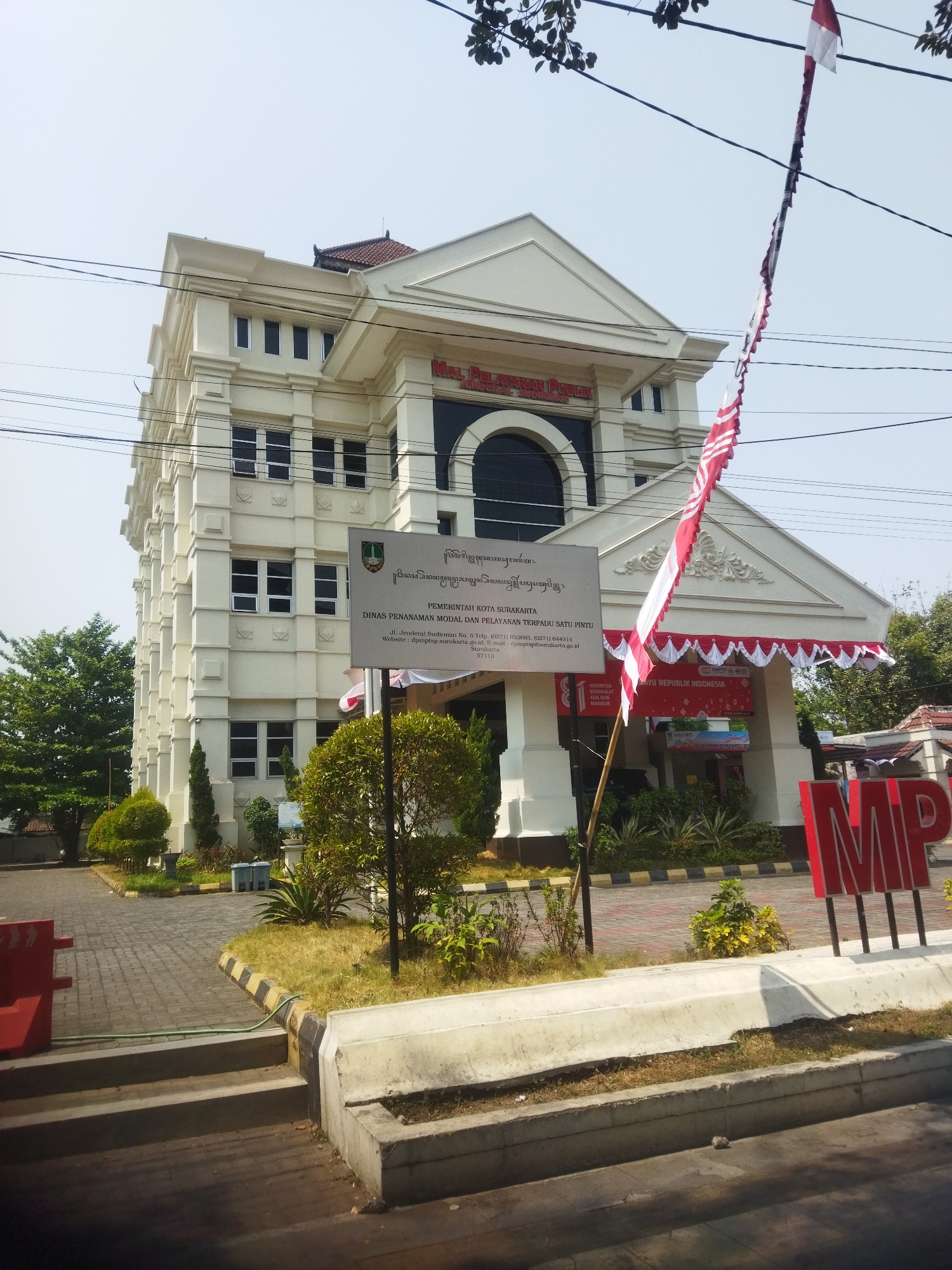 The image captures the exterior of the "Mal Pelayanan Publik" (Public Service Mall) in Surakarta, Indonesia. The building is a multi-story structure with white facade, classical architectural elements, and several windows. A sign in front of the building displays information about the "Pemerintah Kota Surakarta" (Surakarta City Government) and the "Dinas Penanaman Modal dan Pelayanan Terpadu Satu Pintu" (Investment and One-Stop Integrated Service Agency). Indonesian flags are visible, with one being a long banner with a red and white pattern. The surrounding environment includes a paved walkway, greenery, trees, and power lines. A large red sign with the letters "MP" is positioned to the right of the entrance. The scene appears to be an outdoor daytime shot.