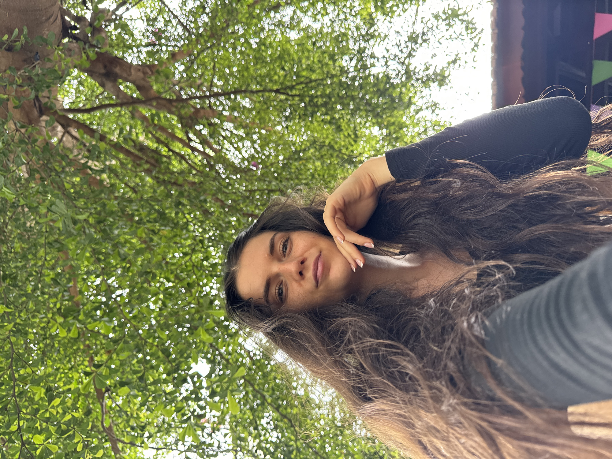 A rotated, low-angle portrait features a young woman with long, dark, wavy hair, looking directly at the camera with a gentle, soft smile. She is wearing a black long-sleeved top, and her right hand, adorned with manicured nails, rests casually against her cheek. Her hair cascades towards the right side of the frame, blending with the background.
The setting is outdoors, beneath a lush canopy of green tree leaves, heavily blurred with prominent bokeh effects from filtered sunlight, indicating bright daytime conditions. Visible tree trunks and branches are interspersed within the foliage. In the upper right quadrant of the background, there's a glimpse of a dark structure, possibly part of a building or a shaded area, and some small, colorful triangular flags or decorations, suggesting a festive or recreational atmosphere. The overall impression is one of natural beauty and tranquility, consistent with an outdoor location like Mũi Né, Vietnam. The woman appears relaxed and content. No text is visible in the image.