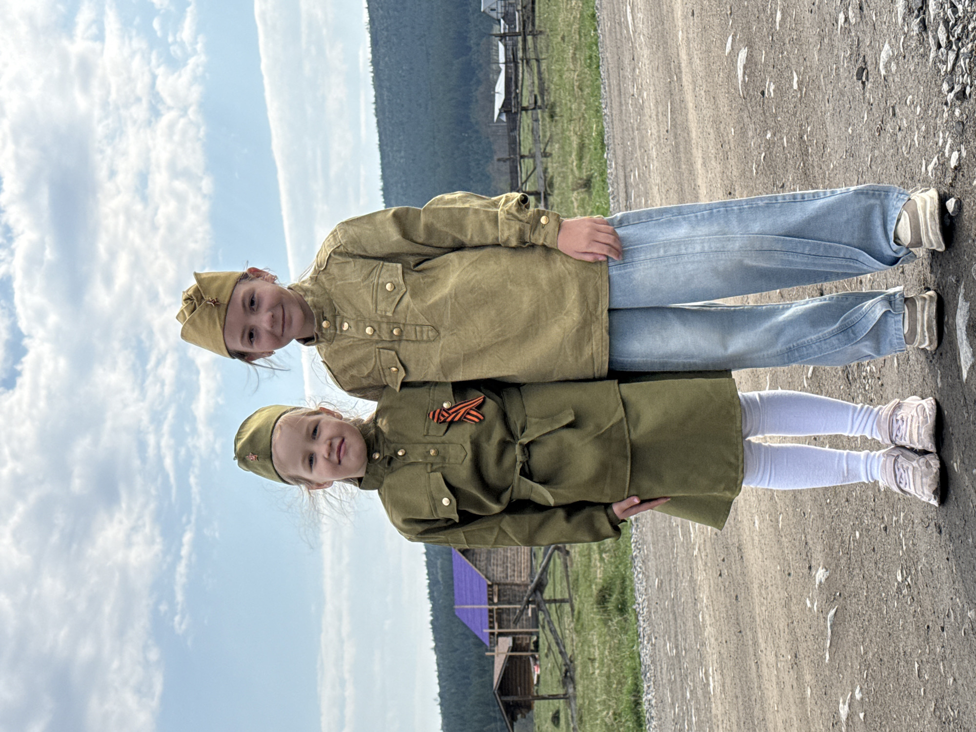 Two young girls stand on a gravel road against a backdrop of a rural landscape. The girl at the top wears a tan military-style shirt, light blue jeans, and sneakers. She also has a military-style cap with a red star. The girl below her wears a green military uniform dress with a belt, white tights, and light-colored sneakers. The girl's dress is decorated with an orange and black ribbon on the chest. She also has a military-style cap with a red star. Behind them is a wooden fence, buildings, and green hills or mountains in the distance. The sky is blue with white clouds. Based on the context of the uniforms, the image is likely related to Victory Day celebrations in Russia. The location is likely Tyulyuk, Russia, due to the style of the buildings.
