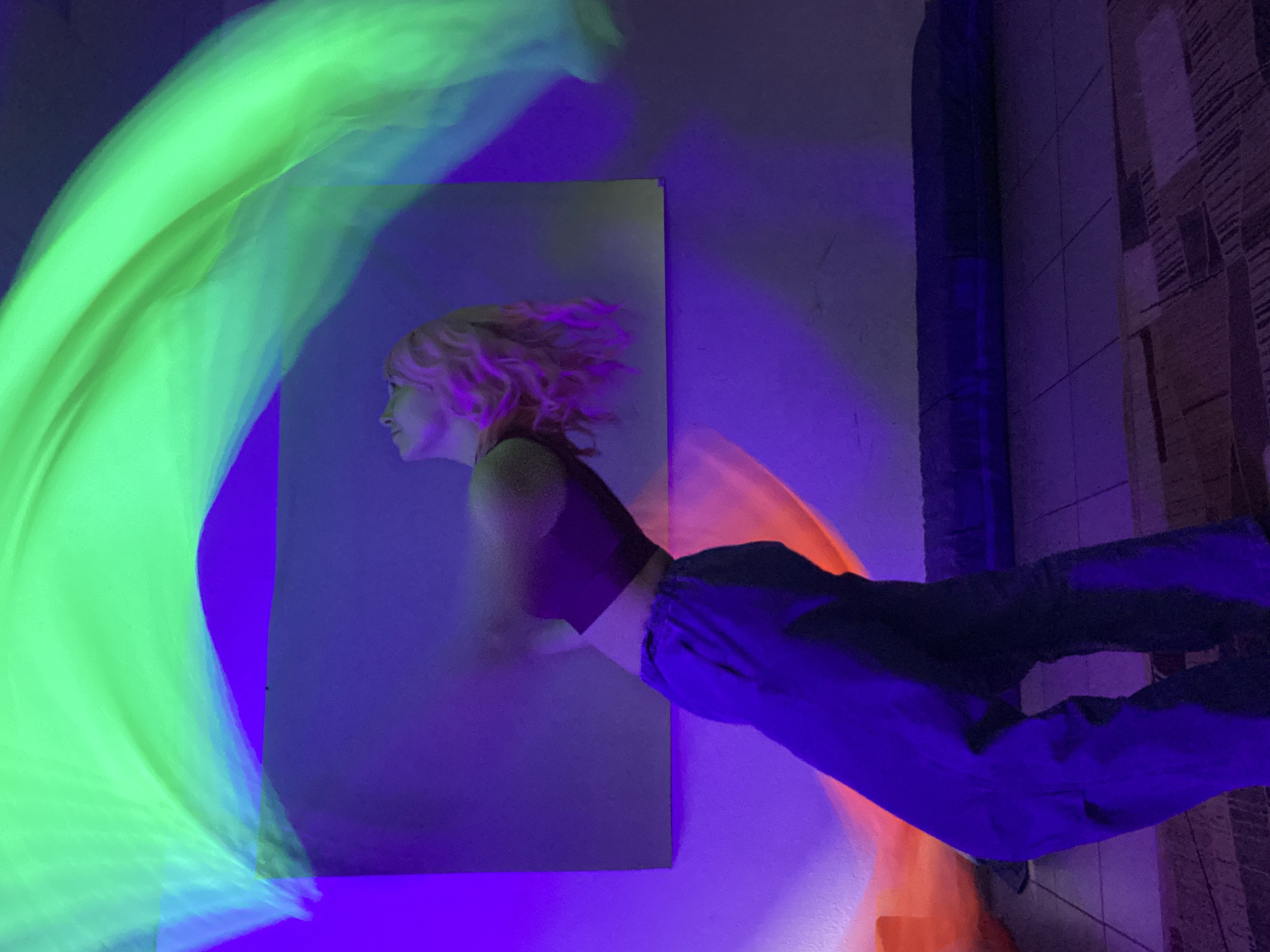 The image shows a brightly lit scene with strong green, blue, and orange lighting effects. A poster or print of a woman with blonde hair, wearing a crop top, is mounted on a wall. A person wearing blue pants is visible, positioned on the right side of the image. A decorative rug or wall covering with a patterned design is on the right side of the image. The location cannot be verified.