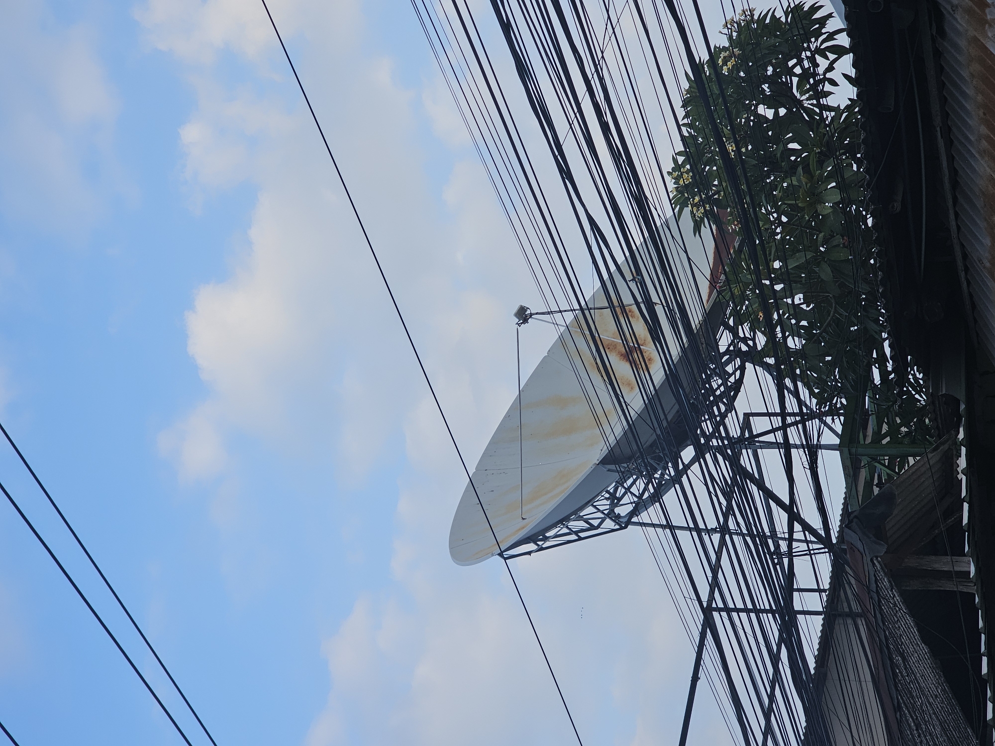 The image, taken in Denpasar, Indonesia, shows a low-angle view of a partially rusted satellite dish mounted on a metal frame, juxtaposed against a bright blue sky with scattered white clouds. A tangle of black electrical wires crisscrosses the scene, partially obscuring the dish and the sky. To the right, the edge of a building with corrugated metal roofing and a tree with white flowers is visible.