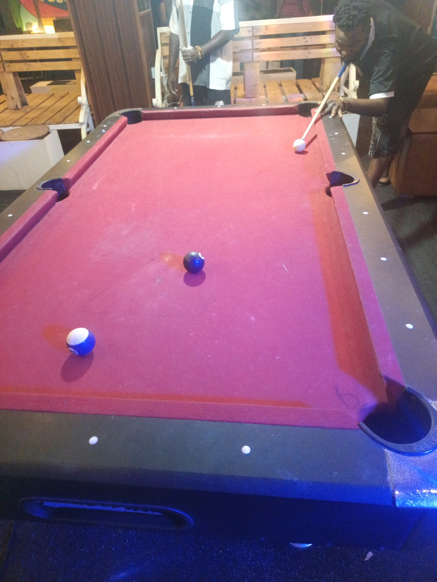 The image depicts a billiards table with a red felt surface. Several pool balls are visible on the table, including a striped blue and white ball and a solid black ball with a white number eight. A white cue ball is positioned near the top of the table, being struck by a pool cue held by a man in a dark shirt. The man is leaning over the table, focusing on the shot. In the background, there are wooden benches and another person standing, partially visible. The scene appears to be an outdoor or semi-outdoor setting, possibly a bar or recreational area. The lighting suggests it is evening or night. There is no specific information in the image to determine the exact location in Obinze, Nigeria.
