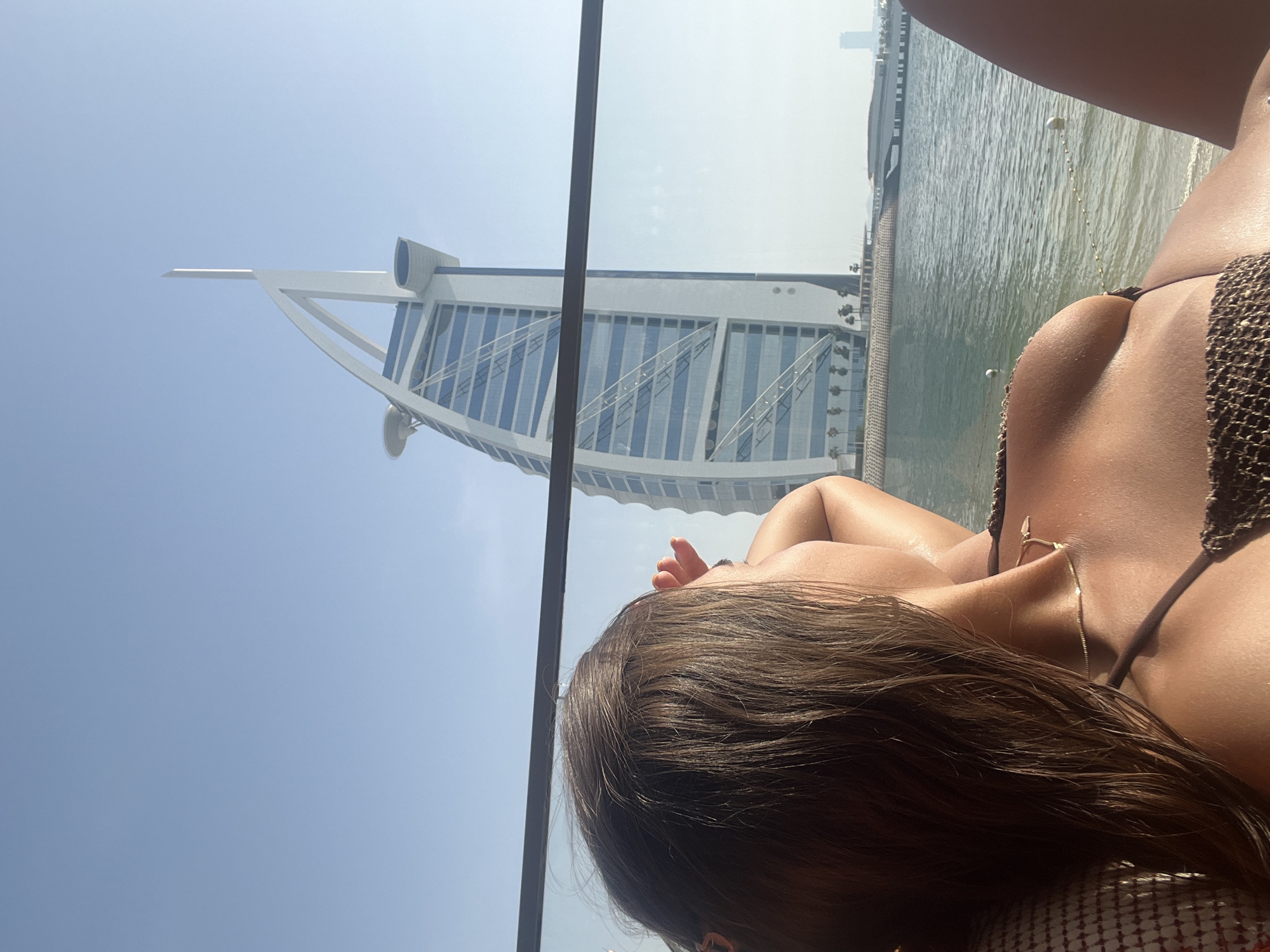 The image is a low-angle perspective photograph of a woman wearing a brown bikini, presumably in Dubai, United Arab Emirates. The Burj Al Arab hotel is visible in the background to the left. In the background, there is a body of water behind the hotel, along with a building further in the distance to the right. There is a structural bar visible in the foreground.