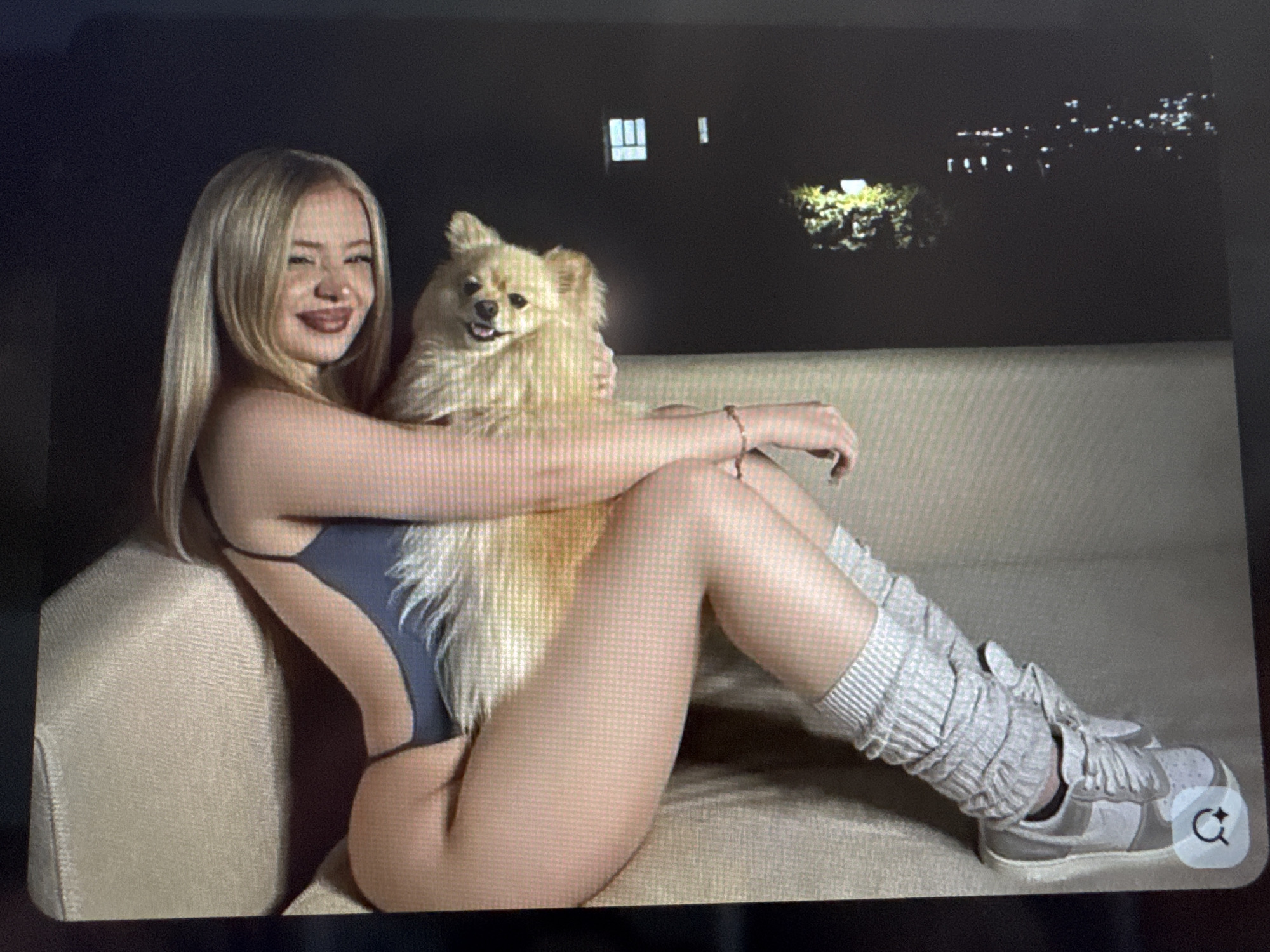 A full-body shot depicts a young woman and a small, fluffy, light-colored dog seated on a light brown couch indoors. The woman has long blonde hair and is positioned on her left side, facing the viewer with a broad smile and dark lipstick. Her left arm is around the dog, which also looks towards the camera with an open-mouthed, happy expression. She wears a dark grey, high-cut, one-piece garment resembling a swimsuit or leotard. Her legs are extended; her left leg is bent, and her right leg is straightened. On her right leg, she wears light grey, ribbed leg warmers over what appears to be fishnet stockings, paired with light grey and white sneakers. A thin bracelet is visible on her right wrist.
The background behind them is dark, indicating nighttime. Through what appears to be a window or open space, distant blurry lights are visible, suggesting a city or residential area at night. Some illuminated foliage is also visible in the middle ground against the dark backdrop. The overall lighting is dim, primarily highlighting the subjects. The image itself appears to be a photograph of a screen, showing slight pixelation. One of the provided crops shows a small, white "magnify/refresh" icon in the bottom right corner of a shoe, which is part of the image viewing interface rather than the original content of the scene.