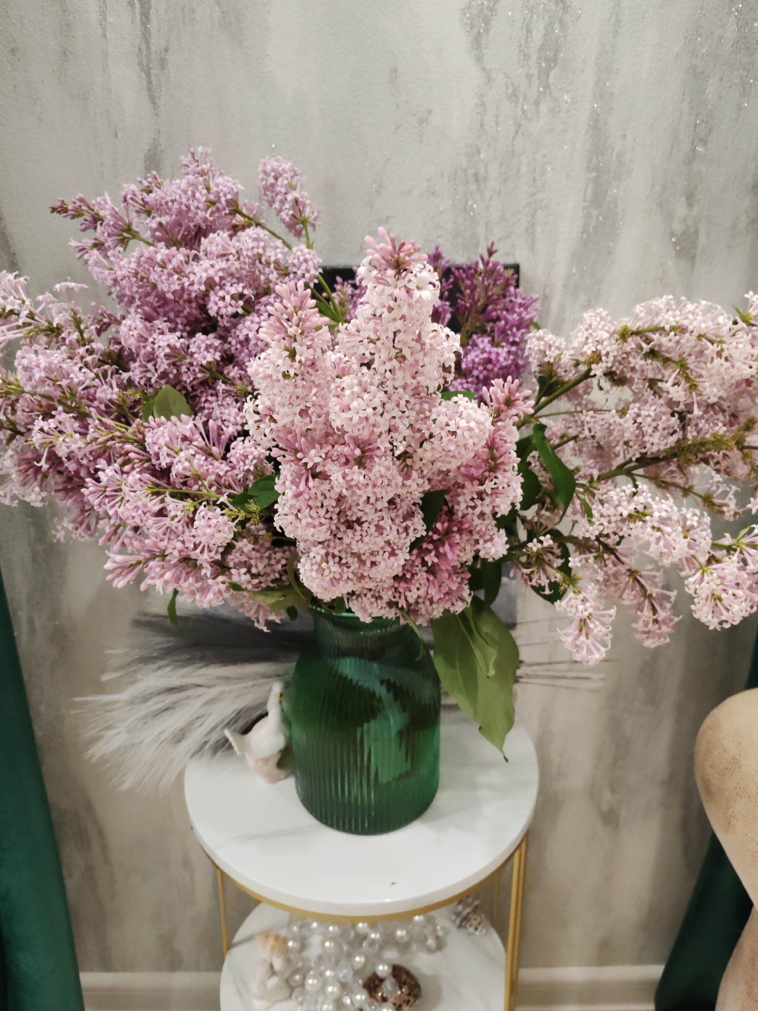 In this close-up, vertical shot, a bouquet of light pink and purple lilacs is arranged in a fluted, emerald green glass vase. The lilacs are abundant, with numerous small florets forming clusters on their stems. Some of the blooms are a soft pink, while others are a deeper, richer purple. A few green leaves are interspersed among the flowers.
The vase sits on a round, white marble-topped table with gold-colored legs. Behind the vase, on the same table, is a fluffy white decoration that resembles a bird or a small animal, and to the left of the vase, a white decorative plume. Below this table, a second, smaller table is visible, adorned with a collection of pearls and shells.
The background is a textured wall with a grey, marbelized pattern and a subtle shimmer. To the left and right of the main subject, dark green velvet curtains are partially visible, suggesting an indoor setting, likely a home or a studio. The lighting appears to be soft and diffused, creating a gentle atmosphere. No people are visible in the image. The overall impression is one of springtime freshness and elegant home decor. The location context of Komsomolsk-on-Amur, Russia, is noted but not visually evident within the frame itself.