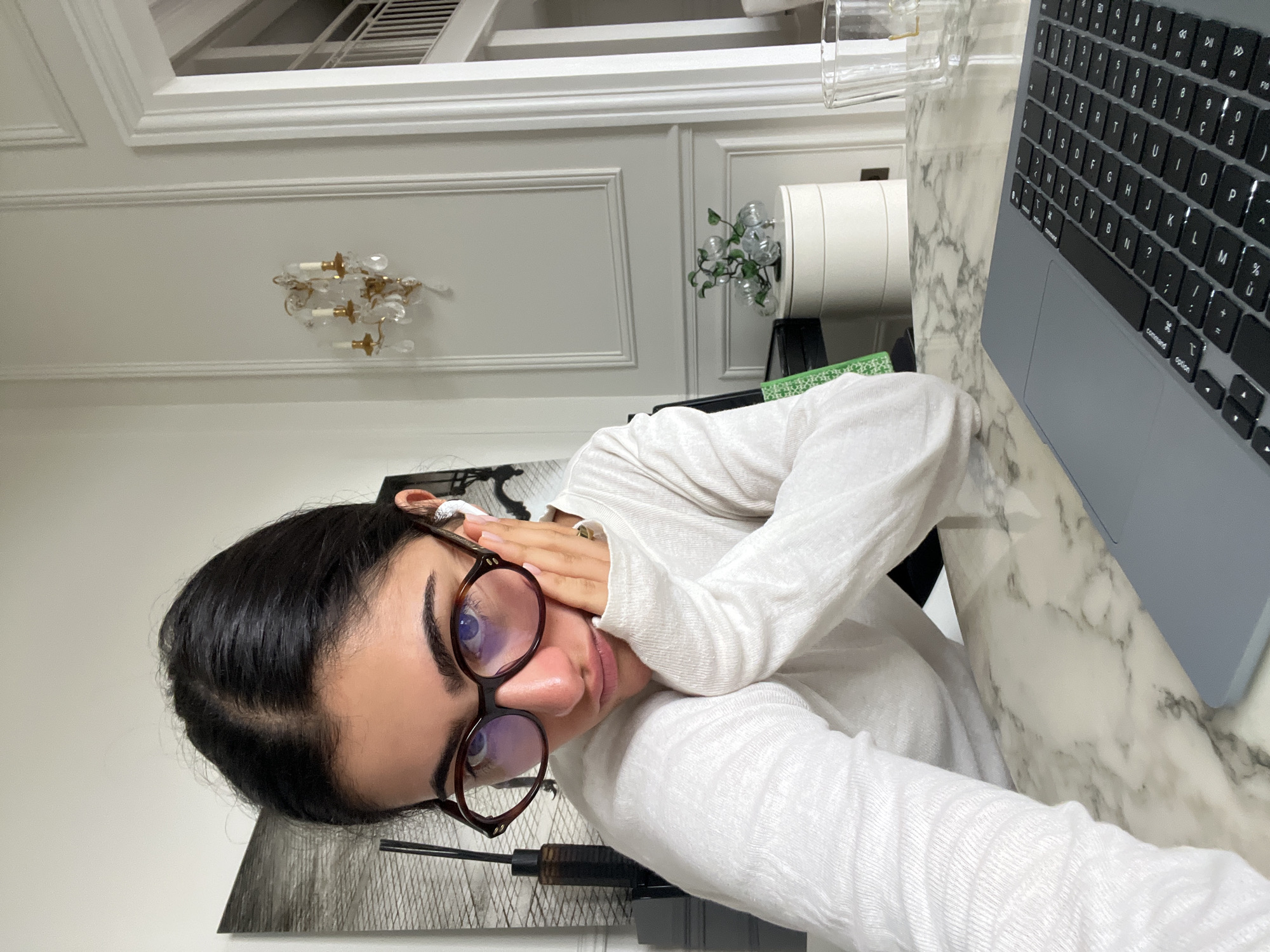 Here is a forensic description of the image:
The image is a close-up, slightly distorted view of a young woman with dark hair, wearing round tortoiseshell glasses and a white long-sleeved shirt. She is positioned at an angle, with her face angled towards the camera and her hand near her face. An earbud can be seen in her ear.
The background includes a laptop with a French keyboard, situated on a marble-like surface. Behind the woman are various decorative elements, including a chandelier, a decorative object resembling plant bulbs in a white pot, and a piece of framed art (possibly an abstract design) leaning against the wall.
Based on the architecture and the style of the interior decor, it is impossible to confirm definitively, but there are no indications that the location is Paris, France. The image is most likely taken indoors, at a residence.
