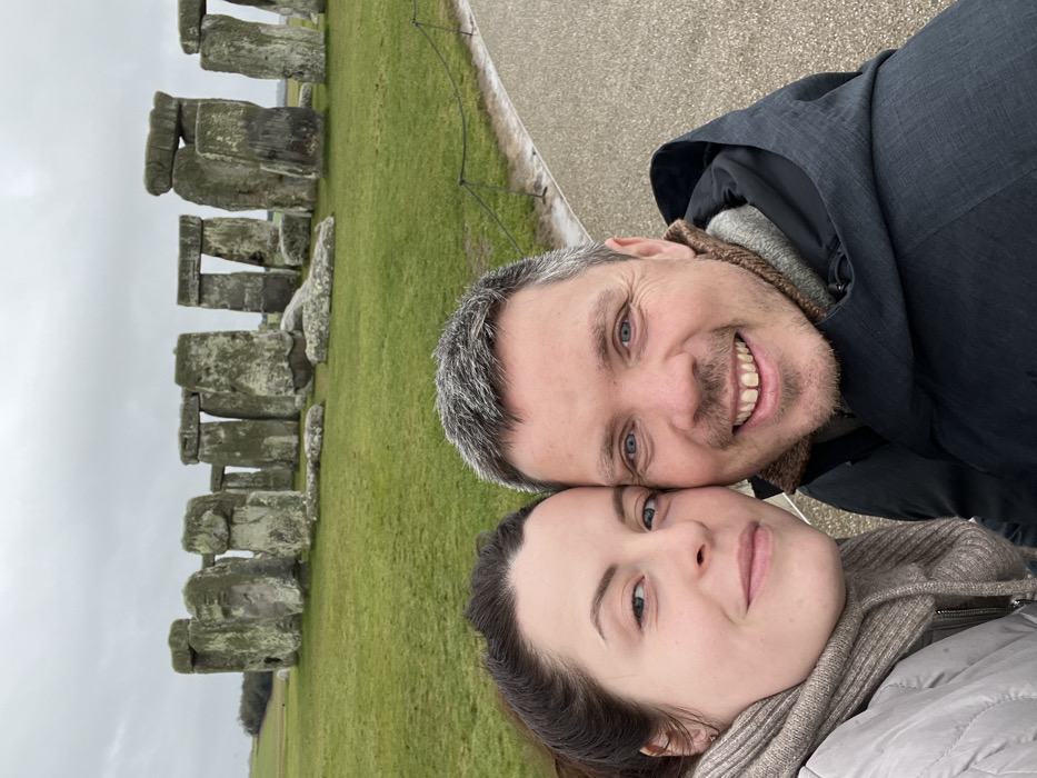 A selfie-style photograph depicts a man and a woman in the foreground, smiling directly at the camera. The man, with short grey and dark hair and blue eyes, wears a dark hooded jacket over a brown fleece-lined collar. He has a gap between his two front teeth. The woman, with dark hair pulled back and blue eyes, wears a light grey puffer jacket and a beige cowl neck scarf. Her face is positioned close to the man's.
In the background, approximately 10-15 meters behind the individuals, stands the ancient monument of Stonehenge. The monument consists of several large sarsen stones arranged in a circular formation, some still upright with lintels connecting them, others partially fallen or in fragments on the green grass. A paved path, visible on the right, curves along the perimeter of the monument, separated by a thin rope barrier supported by black posts. The ground surrounding Stonehenge is covered in vibrant green grass. The sky is overcast and grey. The location is Stonehenge, near the village of Winterbourne Stoke, Wiltshire, United Kingdom.