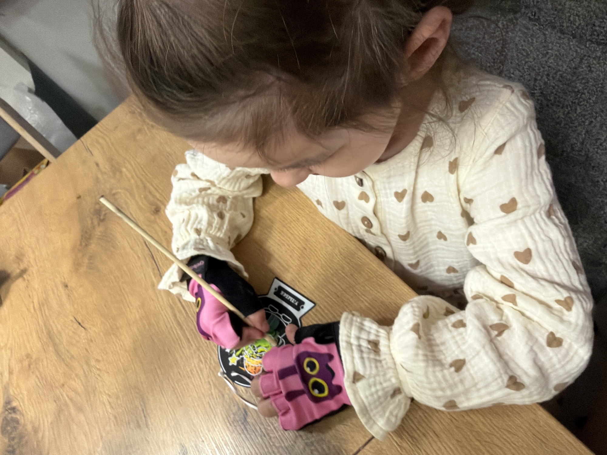 A young child is seated at a wooden table, viewed from above. The child wears a white, heart-patterned long-sleeved shirt and pink fingerless gloves with cartoon faces on the knuckles. The child is using a thin wooden stick to manipulate a black Halloween-themed cutout with graphics of a jack-o'-lantern, stars, and a cauldron. The cutout also has a tag with the word "YMMKA" visible. The location is determined by the tag to be somewhere in the city of Chaikovsky, Russia.