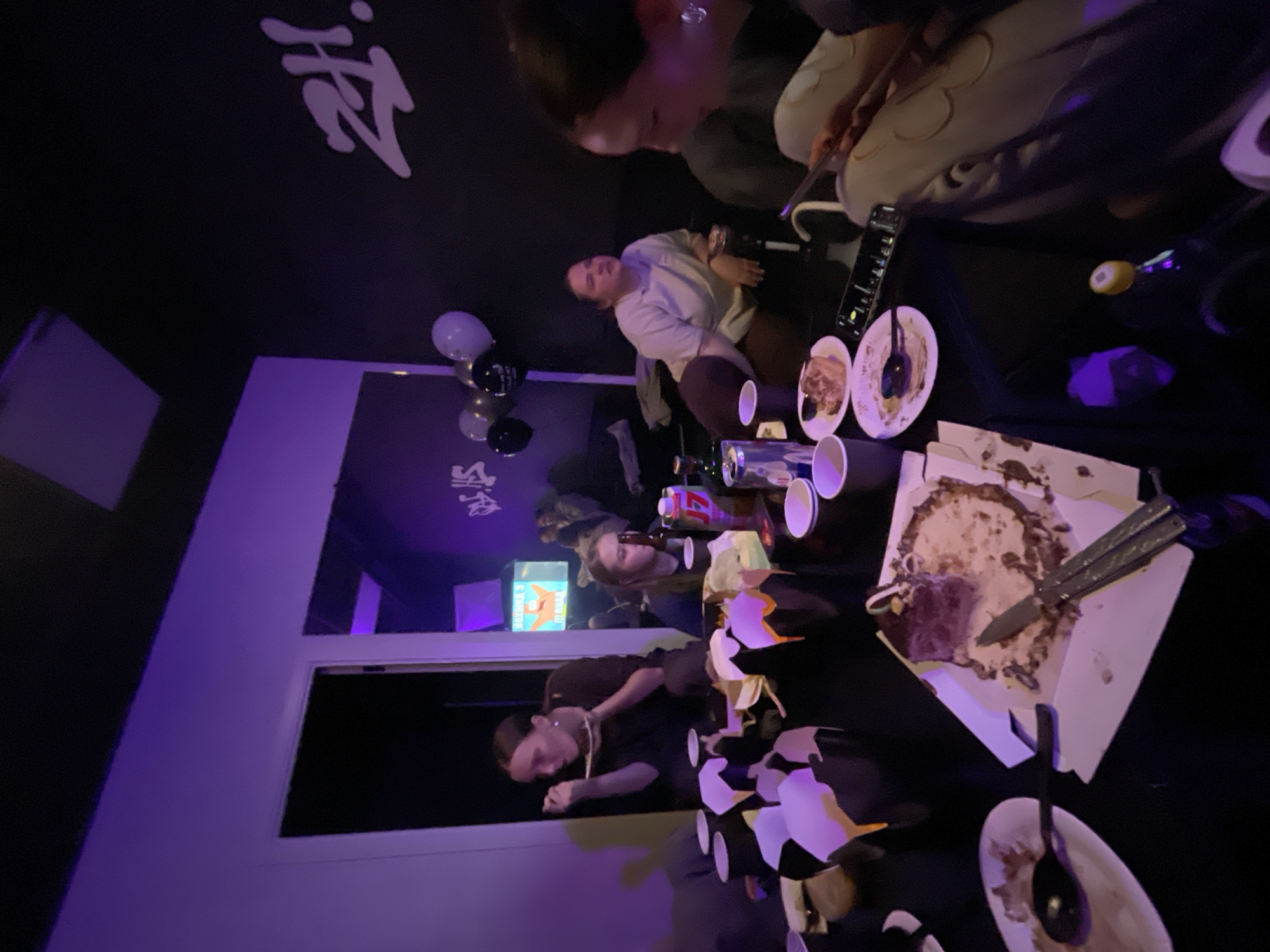 The image depicts an indoor event, likely a party or celebration, with several people present around a table laden with food and remnants of a cake. The setting appears to be a room in the city of Krasnoyarsk, Russia, as evidenced by the presence of a mirror reflecting signage in what appears to be the Russian language. Balloons are visible, suggesting a festive occasion. The table contains disposable cups, plates with leftover food, and a cake box with a partially consumed cake. People are seated and interacting around the table, some using their phones, others engaging in conversation. The room is dimly lit, with purple lighting effects.