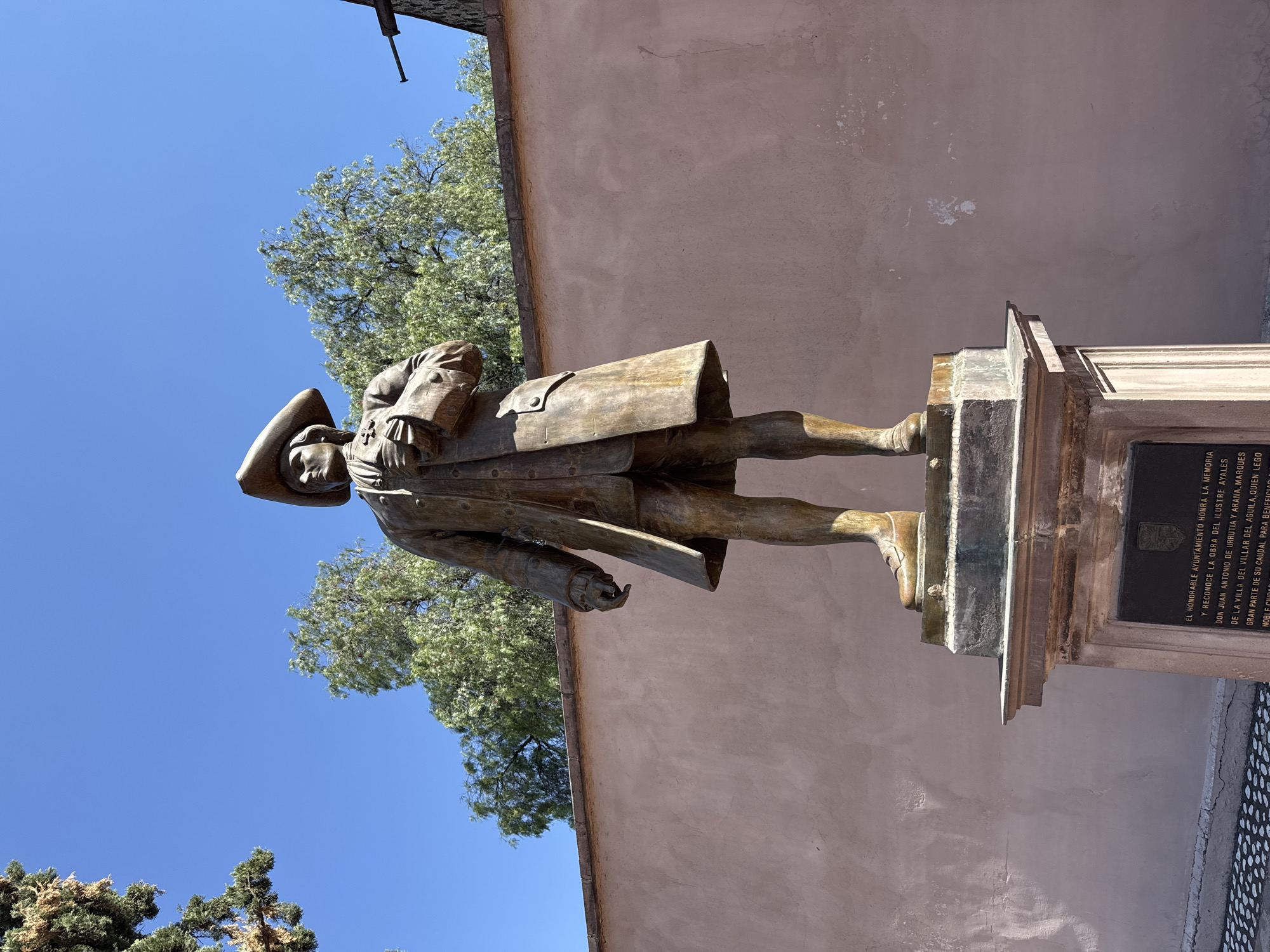 The image depicts a bronze statue of a man in historical attire, including a hat and coat, standing on a stone pedestal. A plaque is affixed to the pedestal. The statue is set against a backdrop of a light pink building and a blue sky with treetops visible. The plaque, written in Spanish, indicates the statue is a tribute to Don Juan Antonio de Urrutia y Arana, Marques de la Villa del Villar del Aguila. The image is likely in the city of Querétaro, Mexico.